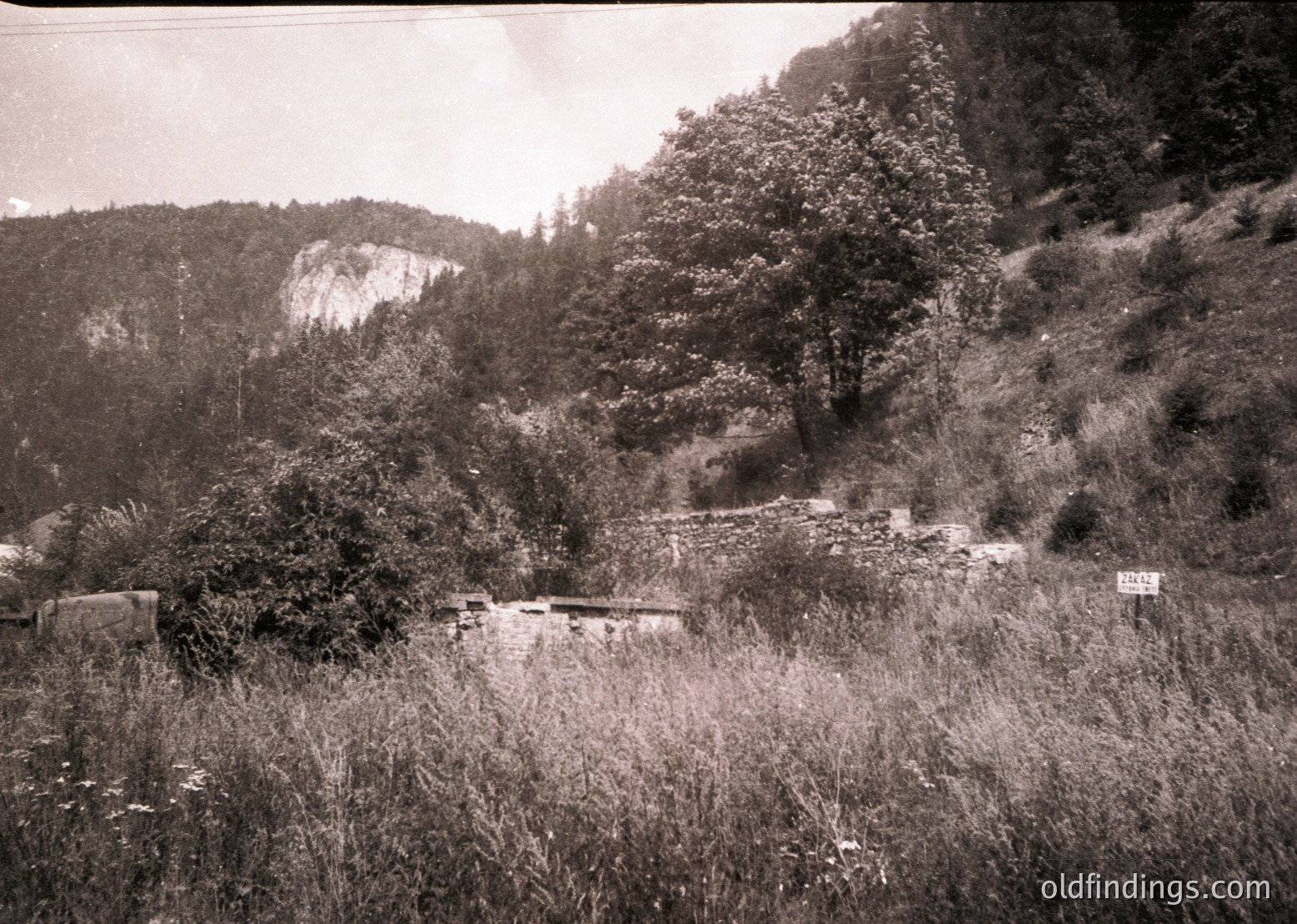 Vintage sepia-toned rural landscape featuring a rocky hillside with sparse vegetation and a narrow, winding dirt path. Foreground includes weathered stone structures, likely bee hives or agricultural storage. Snow-capped mountains in background suggest alpine terrain. Likely early-to-mid 20th century.