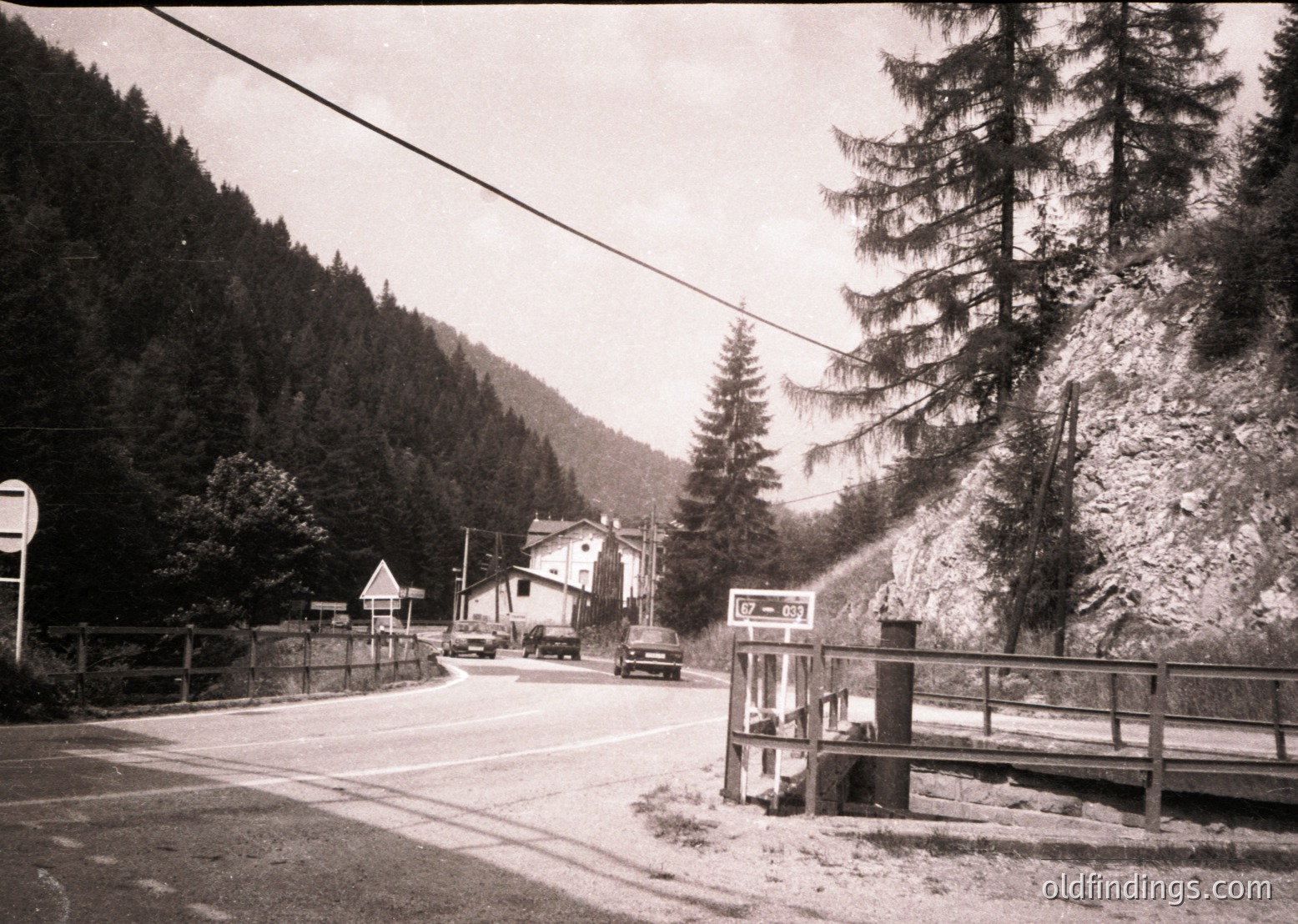 Mid-20th century alpine village roadside with vintage cars, wooden houses, and pine trees. Public phone booth and directional signpost visible.