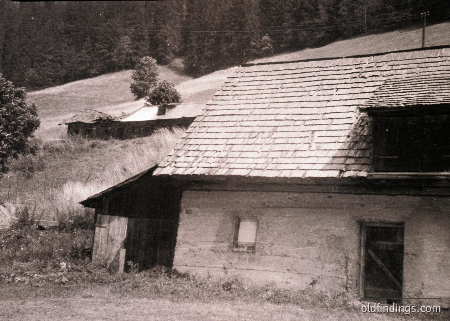 Rustic stone farmhouse with wooden shingle roof, set in a rural valley. Small window and shuttered door suggest mid-20th century construction. Surrounding fields and sparse trees indicate agricultural use. Likely Eastern European countryside, 1950s-1960s.