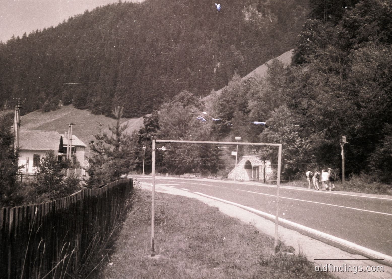 Black-and-white rural roadside scene featuring a single-lane paved road bordered by a wooden fence. A small stone archway marks an entrance to a forested hillside, flanked by dense evergreen trees. A lone house with a pitched roof sits on the left, partially obscured by foliage. The road curves gently into the distance, suggesting a quiet, isolated location. Likely mid-20th century.