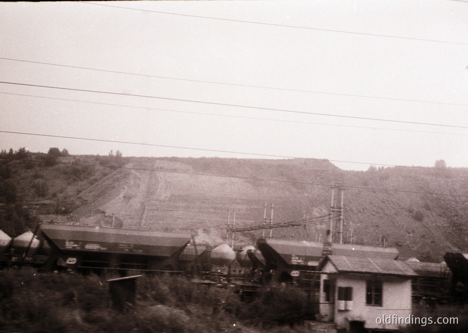 Black-and-white industrial scene featuring a **collapsed railway bridge** with debris scattered across tracks. Foreground shows a small residential building with a pitched roof, likely post-disaster. Overhead, utility poles and wires stretch across a hilly, sparsely vegetated landscape. Suggests **mid-20th century** infrastructure damage, possibly from war or natural disaster.