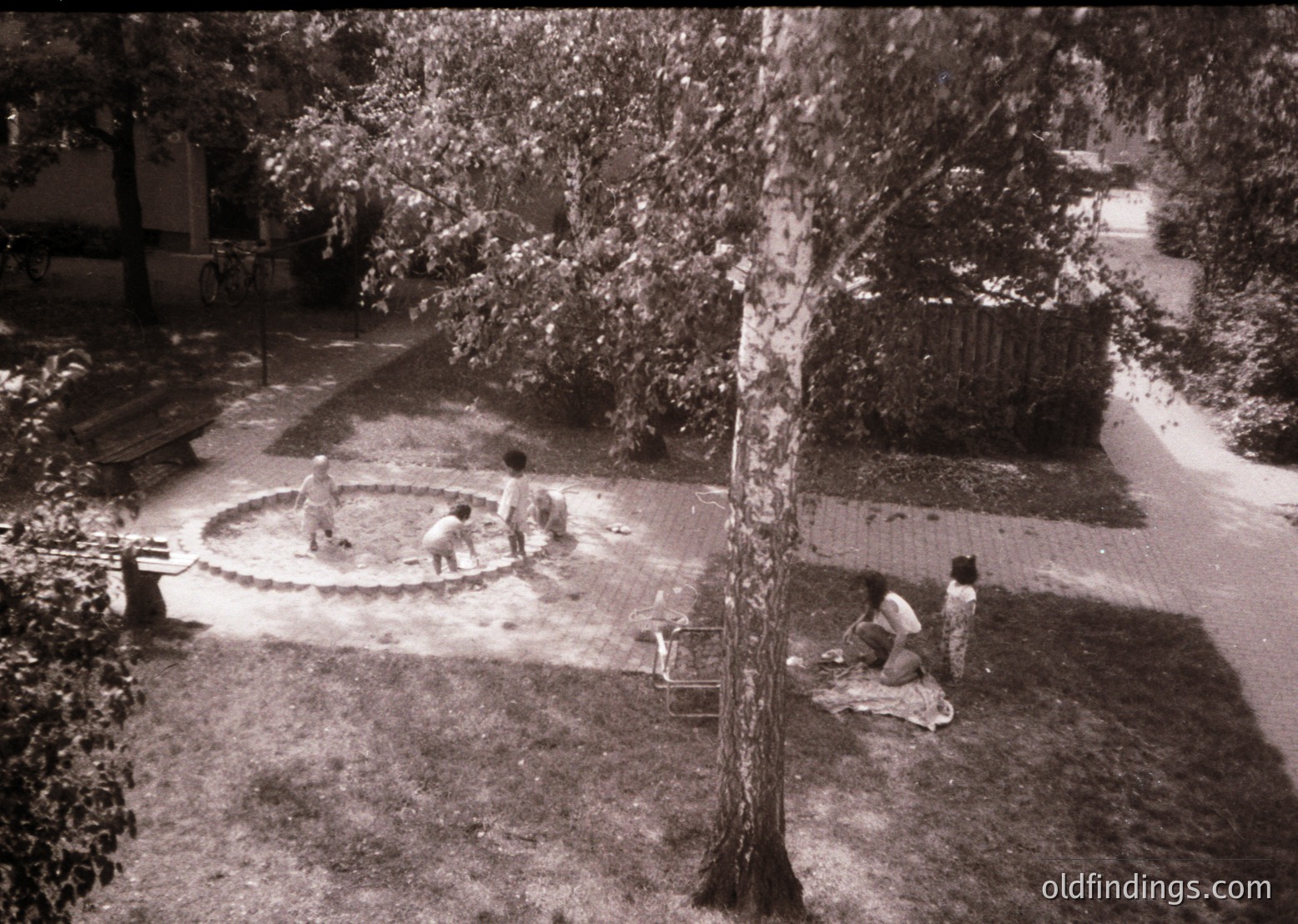 Black-and-white courtyard scene featuring three children playing in a circular concrete water feature. Two children stand at the edge, while one sits on the ground nearby. Surrounding greenery includes a large tree and grassy lawn. Mid-century residential architecture visible in background. Likely 1950s–1970s suburban setting.