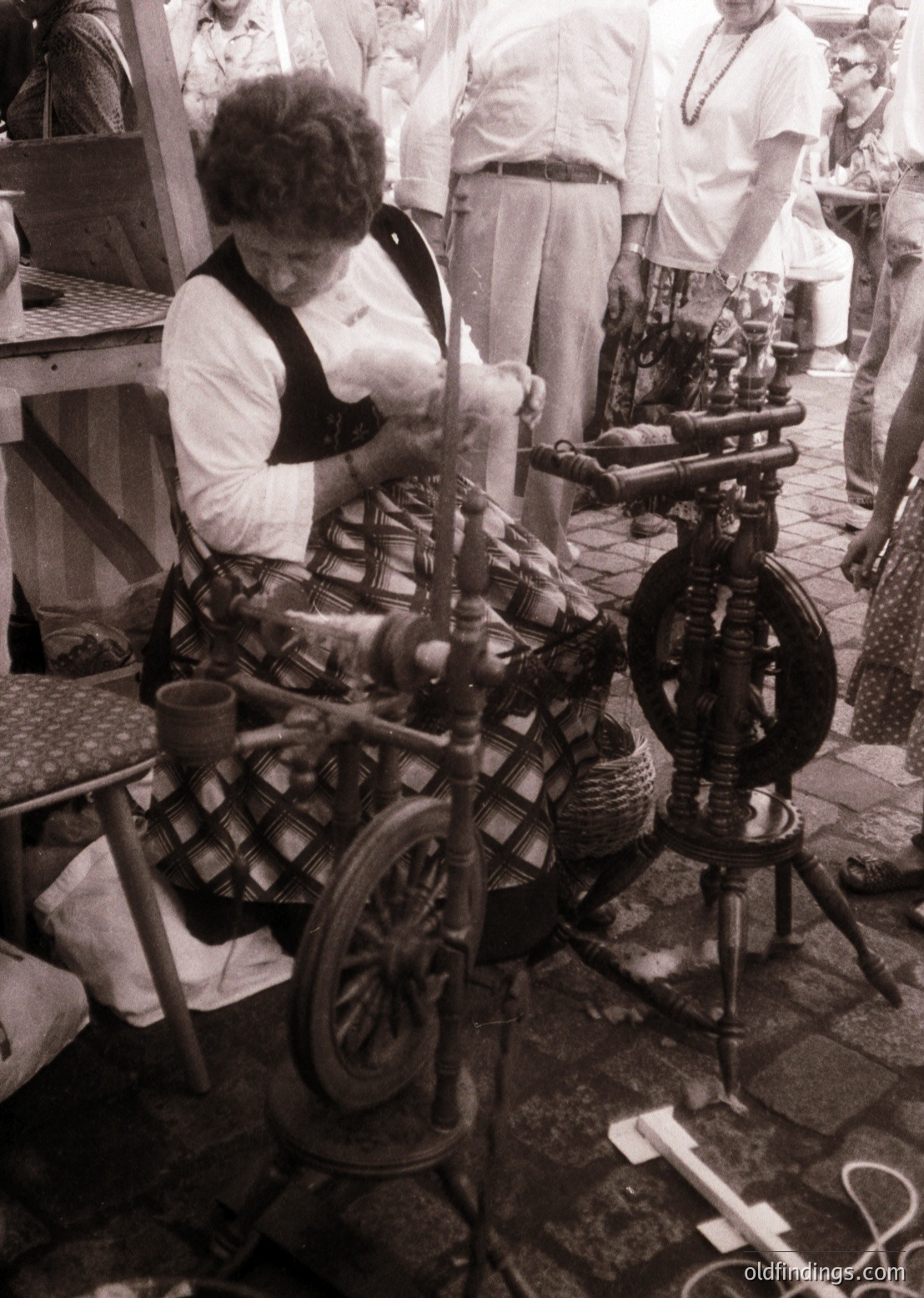 Traditional hand-spinning wheel in use, likely mid-20th century. Woman operates vintage spinning equipment with wool, surrounded by bobbins and woven textiles. Outdoor market or fair setting with blurred spectators observing.