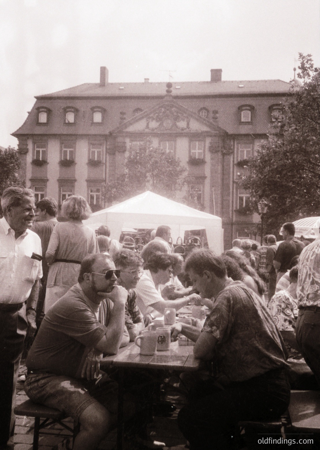 Vintage outdoor gathering at a European café, 1970s. Group of people seated at wooden tables under a white canopy, enjoying drinks and conversation. Neoclassical brick building with decorative cornice in background. Casual summer attire—sunglasses, short sleeves, and hats. Urban European street scene with trees and pedestrians. é