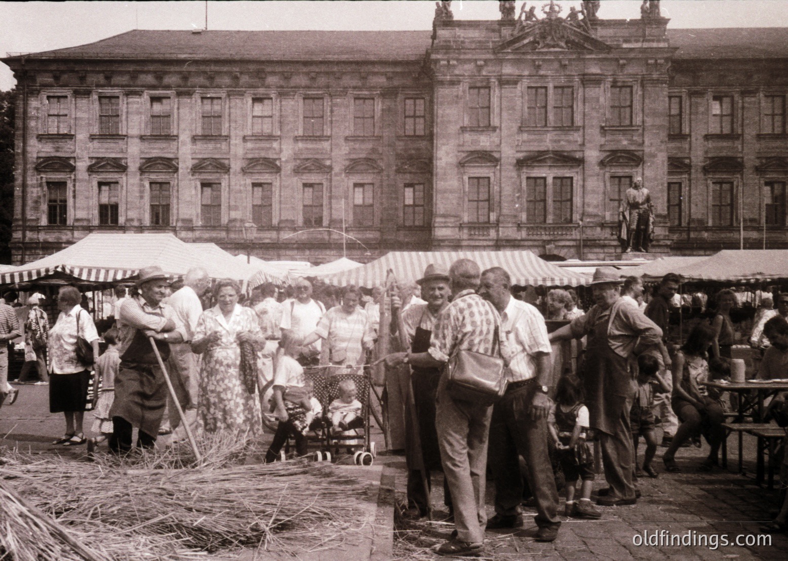 Vintage outdoor market scene featuring a grand neoclassical building with tall columns and arched windows in the background. Crowds gather around stalls covered with white canopies, engaging in transactions. Women in traditional dresses and men in casual 1960s attire interact, with a child in a stroller. Hay bales and rustic elements suggest a rural or semi-urban setting.