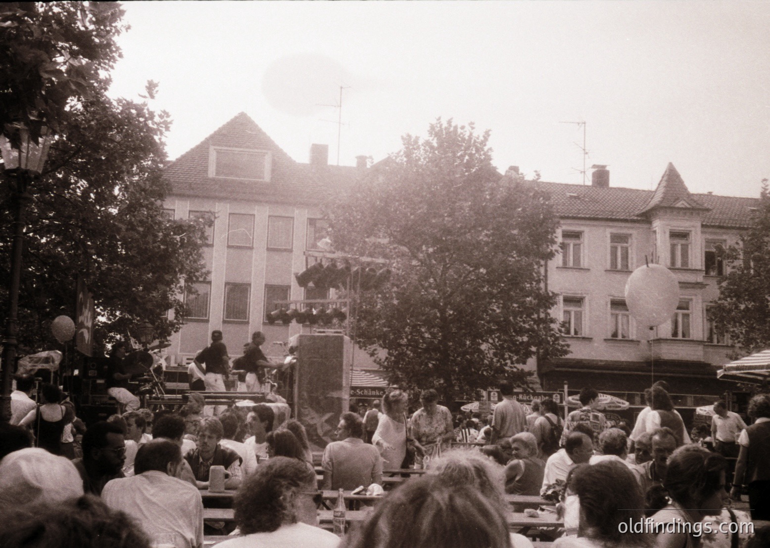 Black-and-white street scene featuring a lively outdoor gathering in a European town square, likely 1960s–1970s. Crowd seated at wooden tables under trees, with a stage elevated on scaffolding hosting performers. Nearby buildings exhibit classic brick facades and gabled roofs. Atmosphere suggests a community festival or public event.