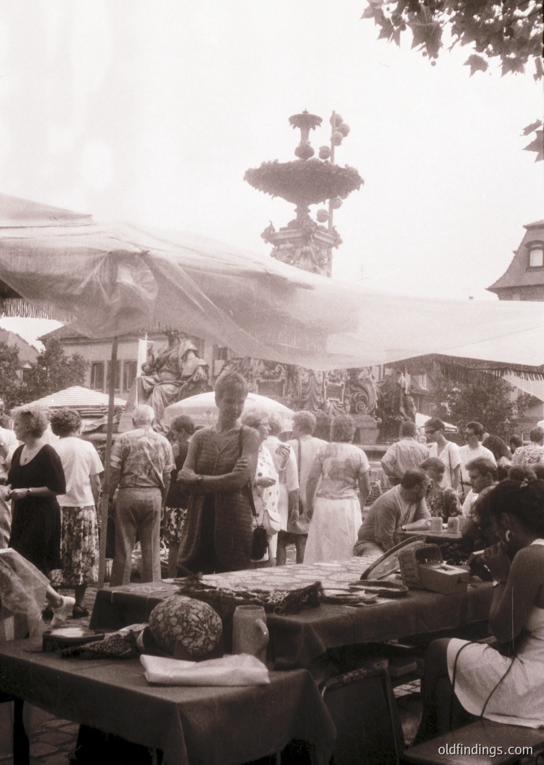 Vintage outdoor market scene featuring a bustling crowd around handcrafted food stalls. Ornate fountain with intricate ironwork dominates the background. Clothing suggests mid-20th century (1950s–1960s).