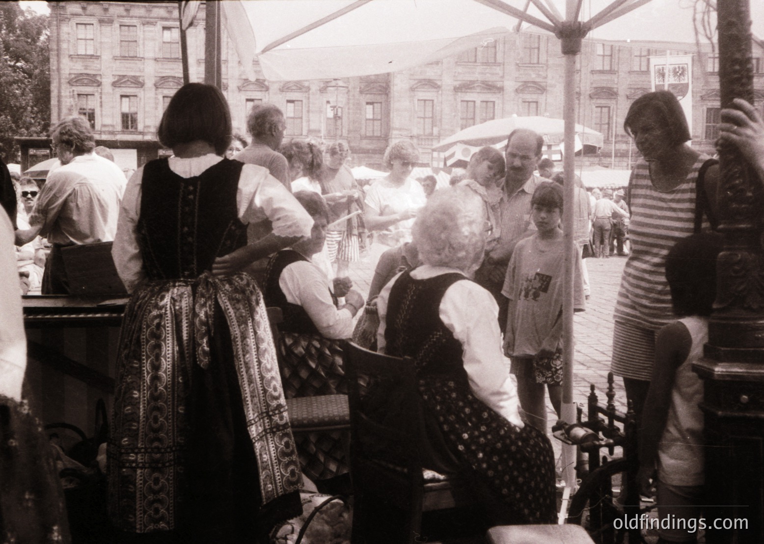 Vintage black-and-white street scene featuring a lively outdoor gathering, likely a folk festival or market. Women in traditional embroidered dresses and vests engage with visitors, showcasing handcrafted items. Men in casual 1970s attire observe or interact. Umbrellas and historic European architecture (brick buildings, arched windows) suggest a European setting. Crowd composition hints at community cultural preservation.