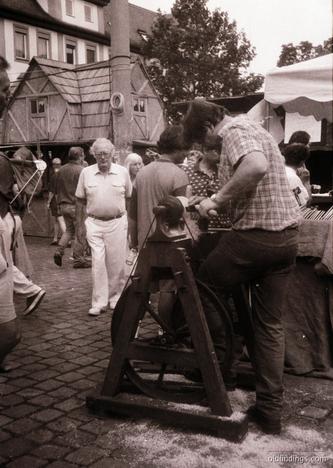 Vintage black-and-white street scene featuring a man operating a manual spinning wheel, likely for wool or textile demonstration. Surrounding crowd in casual 1960s–70s attire, including a man in a white shirt and shorts observing. Wooden barn-style structure and cobblestone pavement suggest a rural or heritage fair setting. Authentic craftsmanship and folk traditions highlighted.