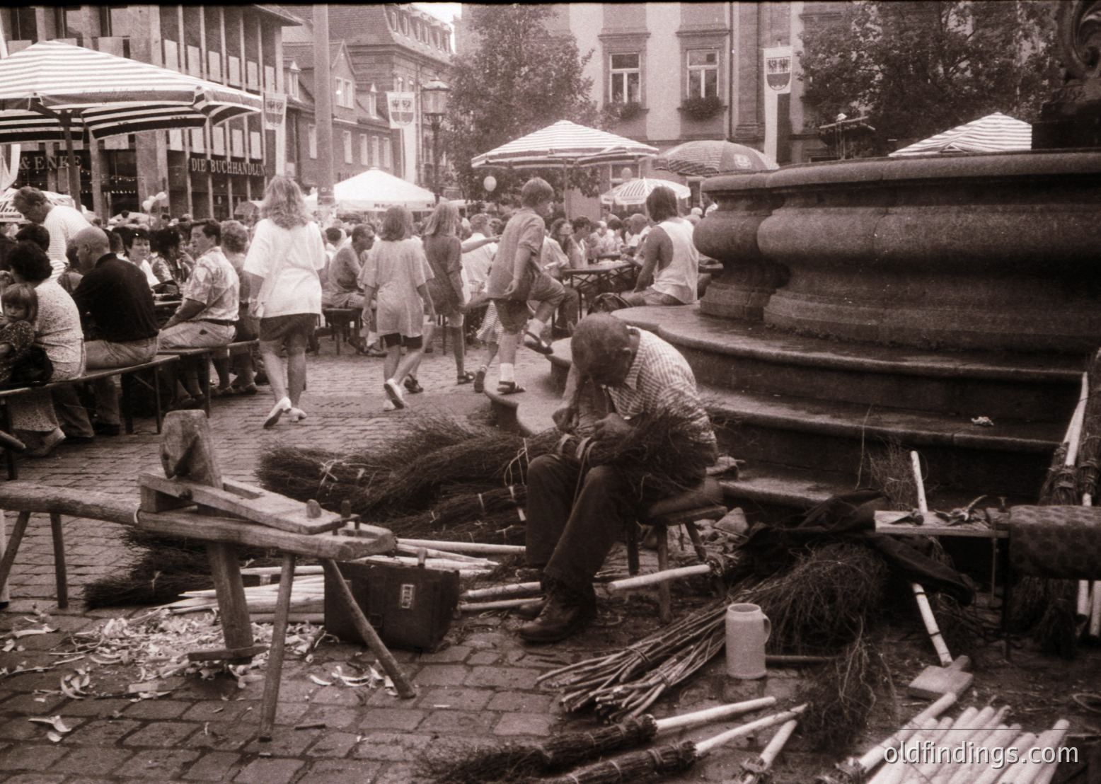 Street vendor sharpening tools beside cobblestone plaza. Mid-20th century European café scene with umbrellas and outdoor seating. Artisan work contrasts with leisurely crowd.