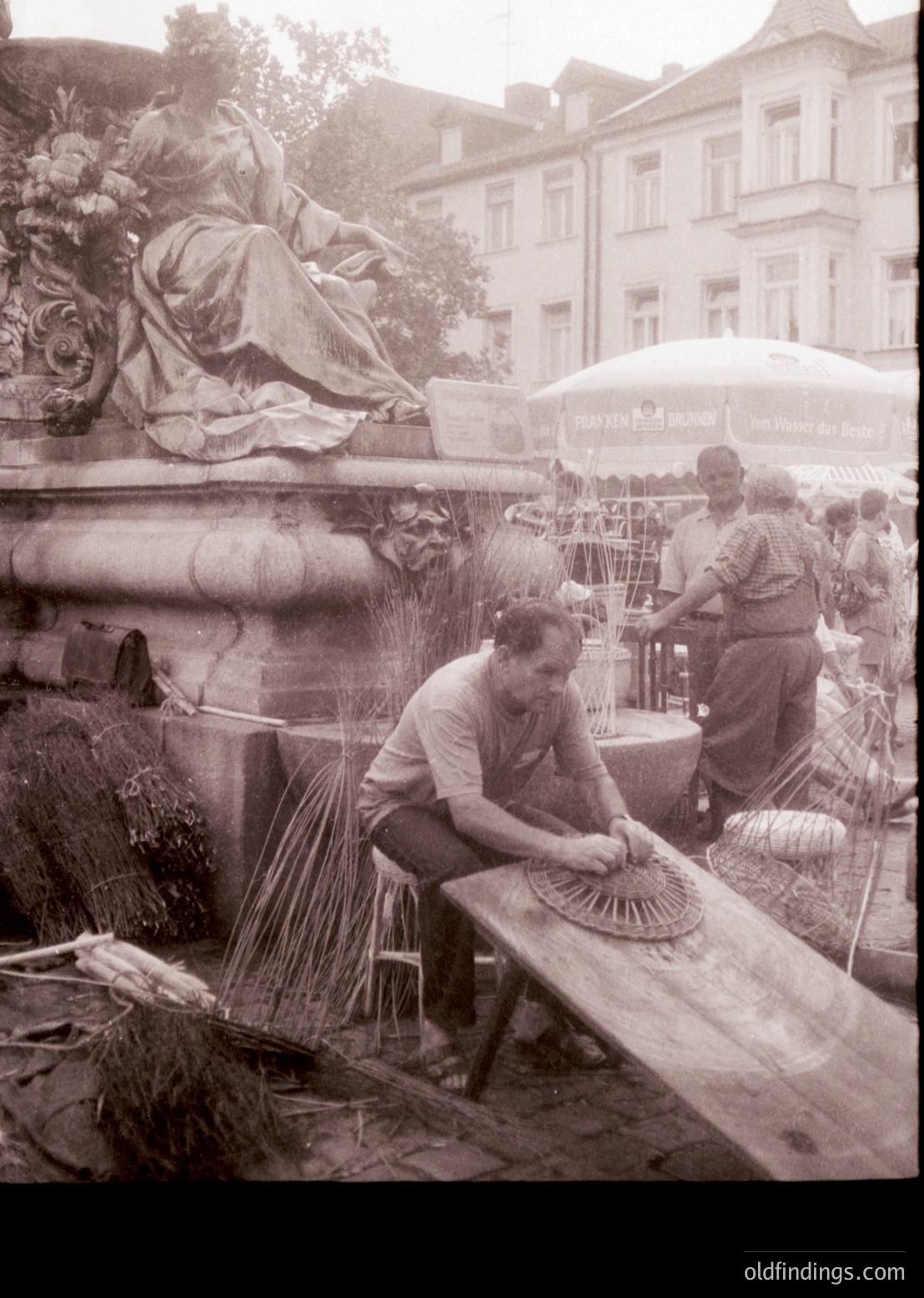 Vintage black-and-white street scene featuring two artisans crafting traditional straw hats on a wooden bench beside a classical stone fountain. Surrounding buildings exhibit early 20th-century European architecture. Crowds and market stalls suggest a lively public square.