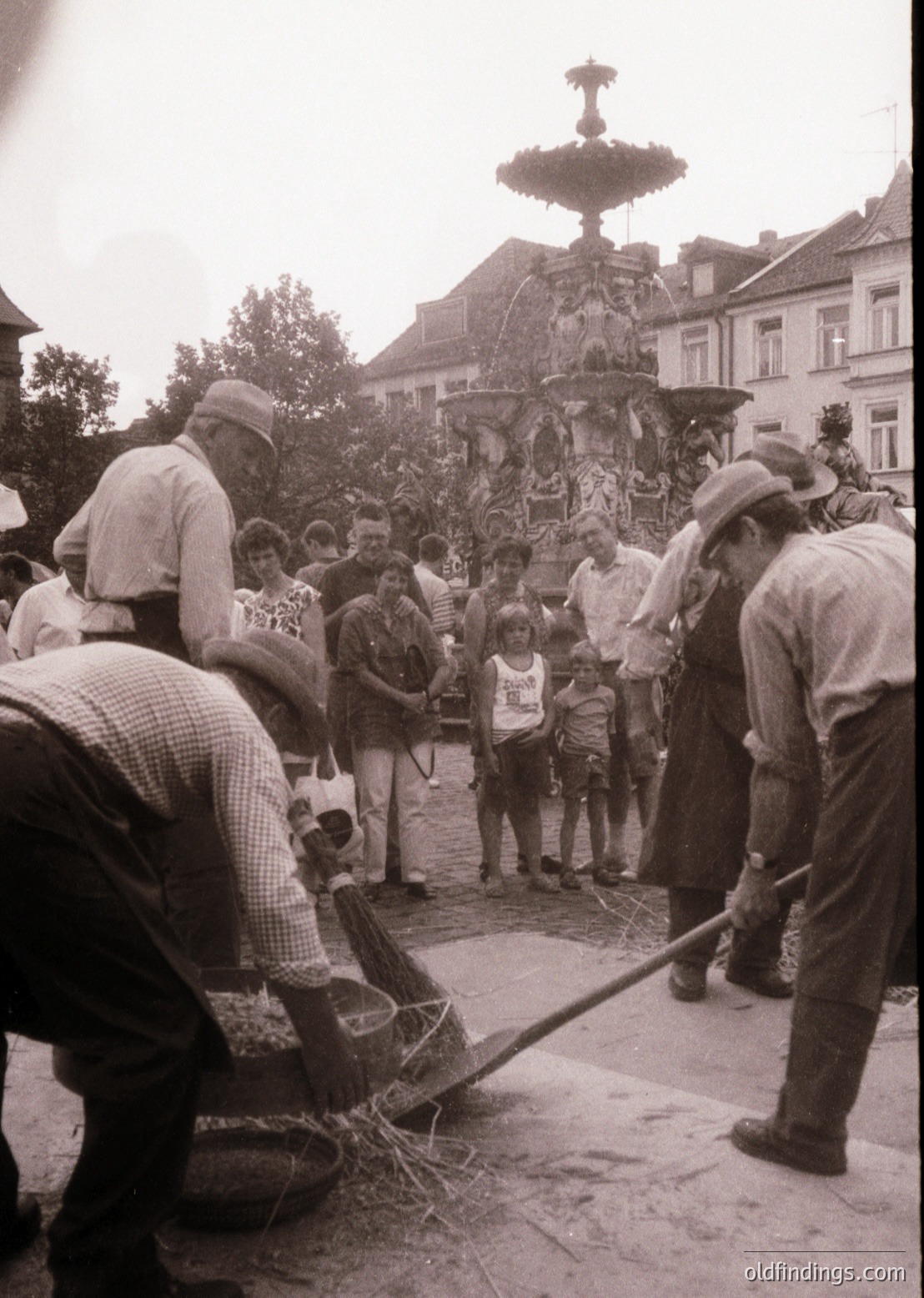 Vintage street scene featuring a group of men clearing debris around an ornate, multi-tiered fountain in a European town square. Mid-20th century clothing (1950s–1960s) and architecture suggest a post-war European setting. Crowd includes onlookers, likely locals, observing the cleanup effort.
