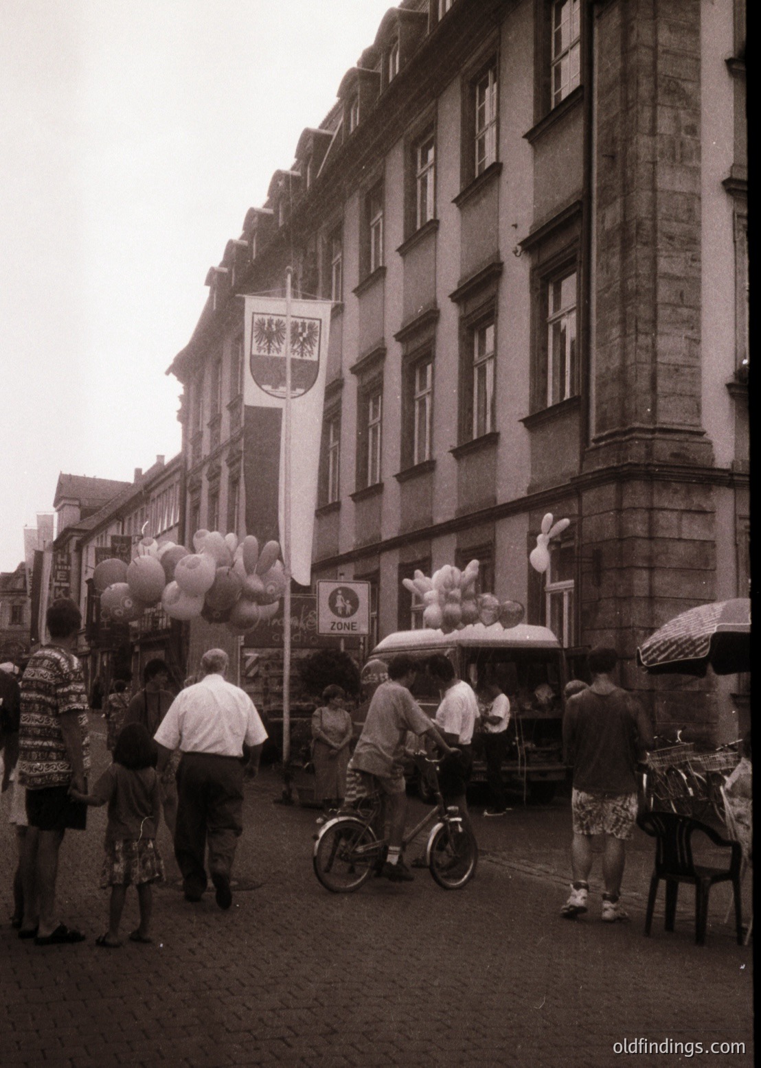 Mid-20th century European street scene with vintage charm. Crowd gathers around a balloon vendor under a historic building featuring a crest banner. Cyclists and pedestrians in casual 1960s attire. Stone architecture with large windows and signage for "Egon" store.