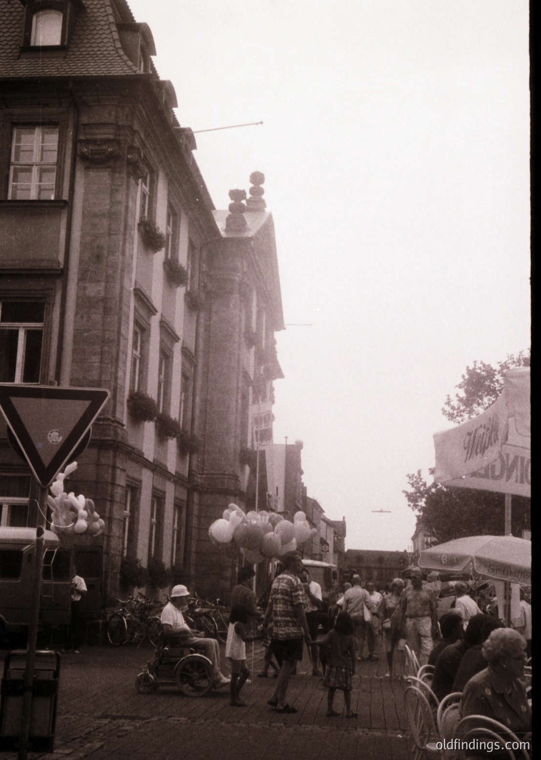 Historic European street scene with mid-20th century architecture—multi-story brick buildings with ornate gables and wrought-iron balconies. Crowds gather under misty skies, likely a market or fair, with balloon vendors and bicycles. Signage hints at a European locale (, , , , ).