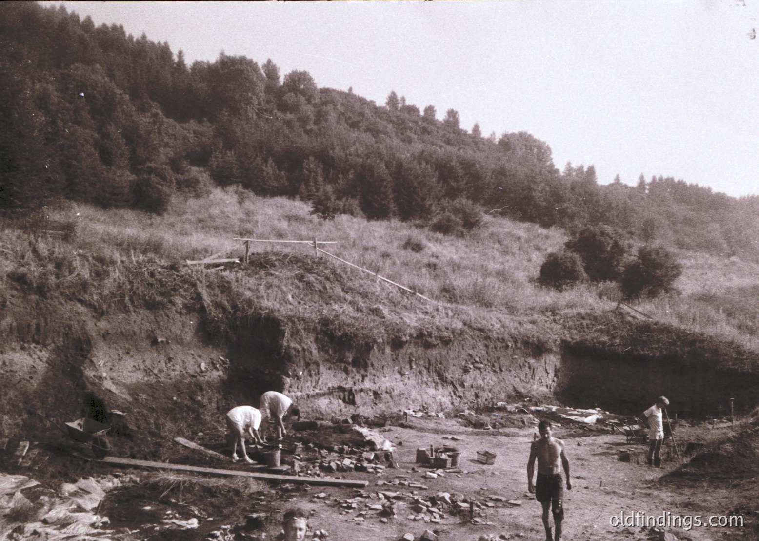 Black-and-white photo of early 20th-century tunnel construction in a forested mountainous region. Workers in period attire use manual tools to excavate a narrow passage; debris and equipment scatter the site. Lush greenery and rocky terrain frame the scene, suggesting rugged terrain. Ideal for historical engineering studies or vintage industrial archives.