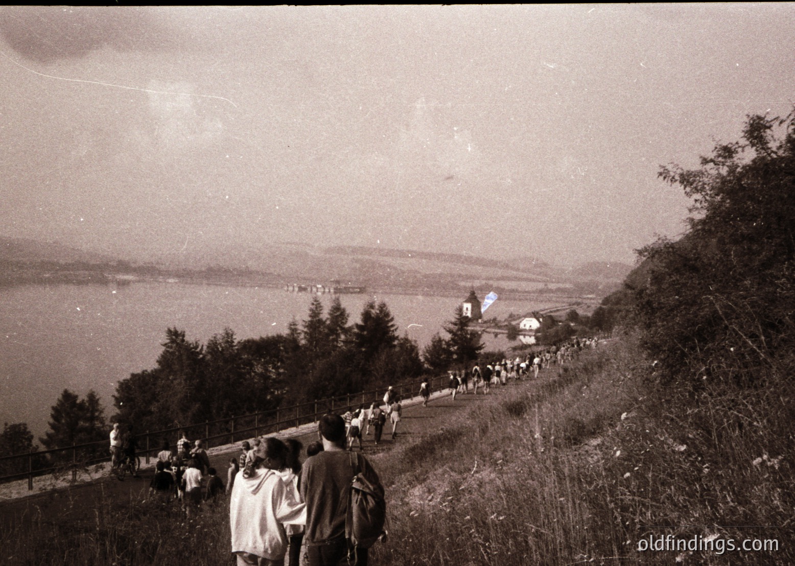 Black-and-white shot of a coastal hillside path crowded with mid-20th-century attire (1950s–60s). Group of hikers descends toward a lakeside chapel and scattered buildings. Dense forest frames the path, leading to a serene waterfront vista.