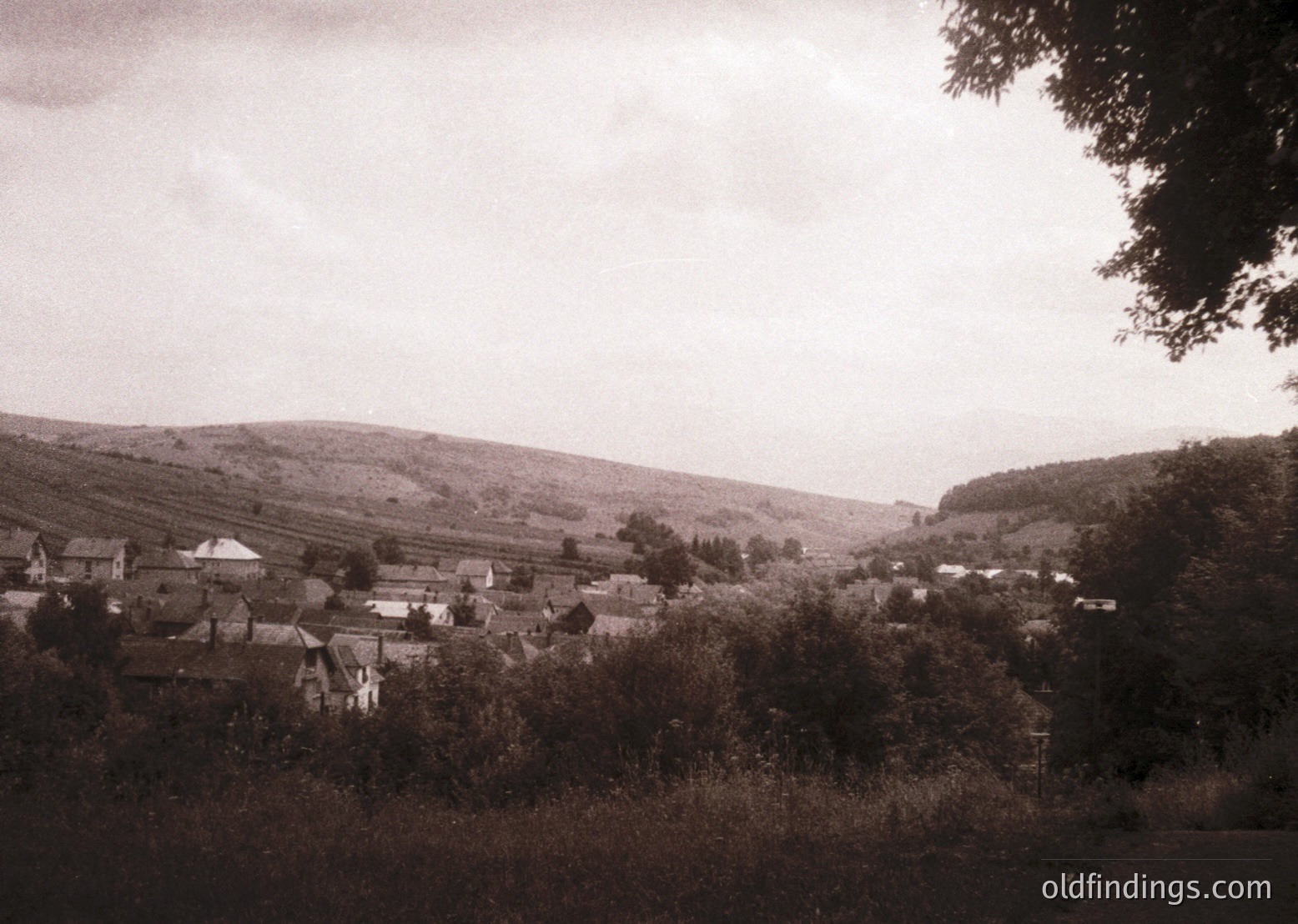 Vintage sepia-toned rural village nestled in rolling hills, likely Eastern Europe. Clustered brick homes with pitched roofs and small gardens, framed by dense foliage and a winding dirt path. Overcast sky enhances nostalgic atmosphere.