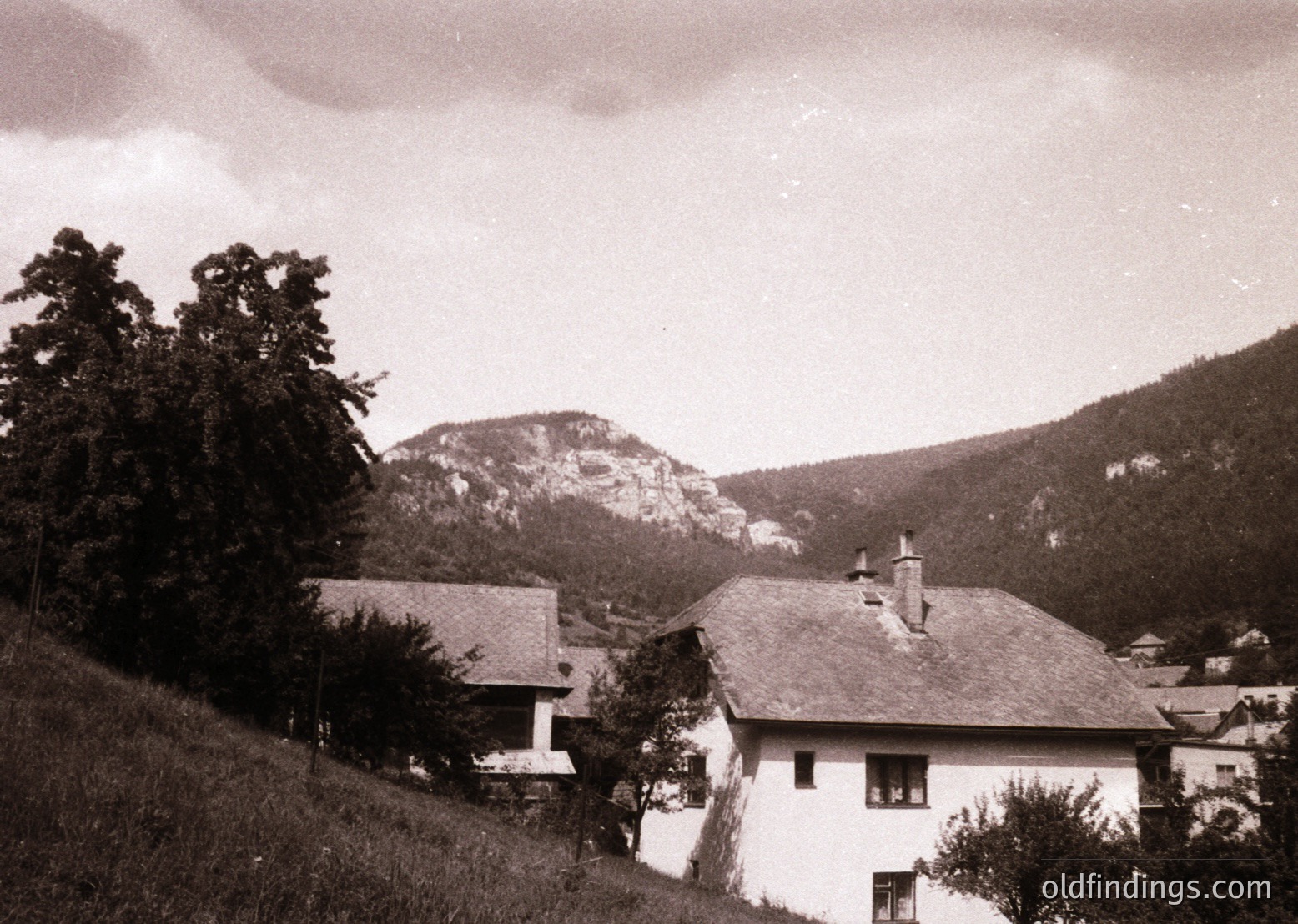 Vintage alpine village scene with rustic wooden houses nestled in a valley, surrounded by forested hills and rocky peaks. Mid-20th century architecture reflects traditional European mountain design.
