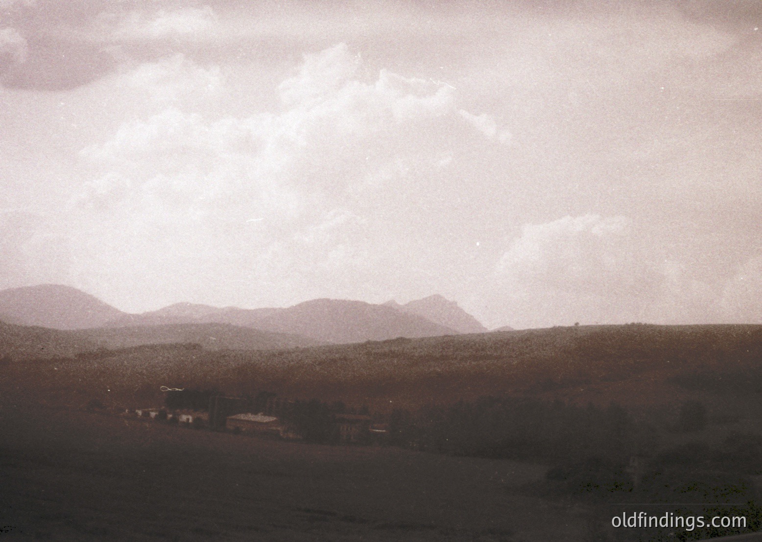 Vintage sepia-toned landscape featuring rolling hills under dramatic, textured clouds. Distant structures suggest rural settlement, likely mid-20th century. Soft focus enhances nostalgic, timeless quality.