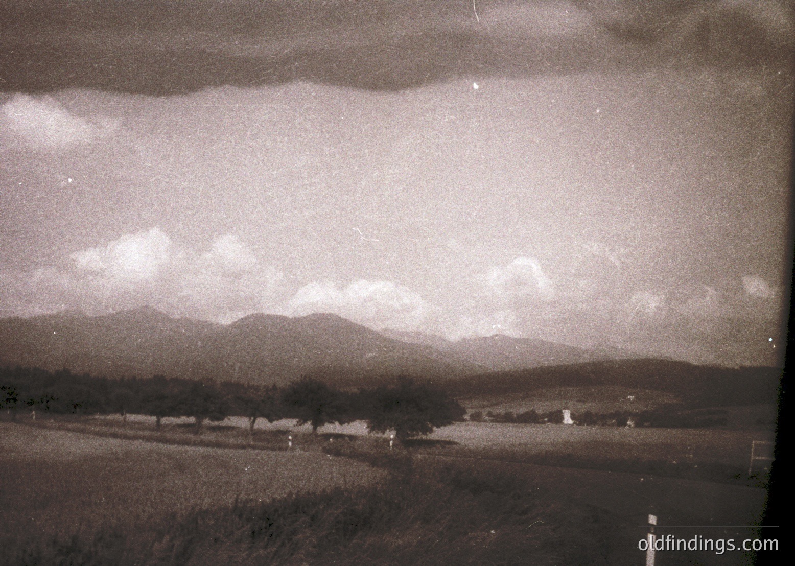 Vintage sepia-toned landscape featuring rolling hills, sparse trees, and a winding road under a dramatic sky. Low-contrast grain suggests early 20th-century photography. Rural setting with minimal human presence, evoking nostalgia.