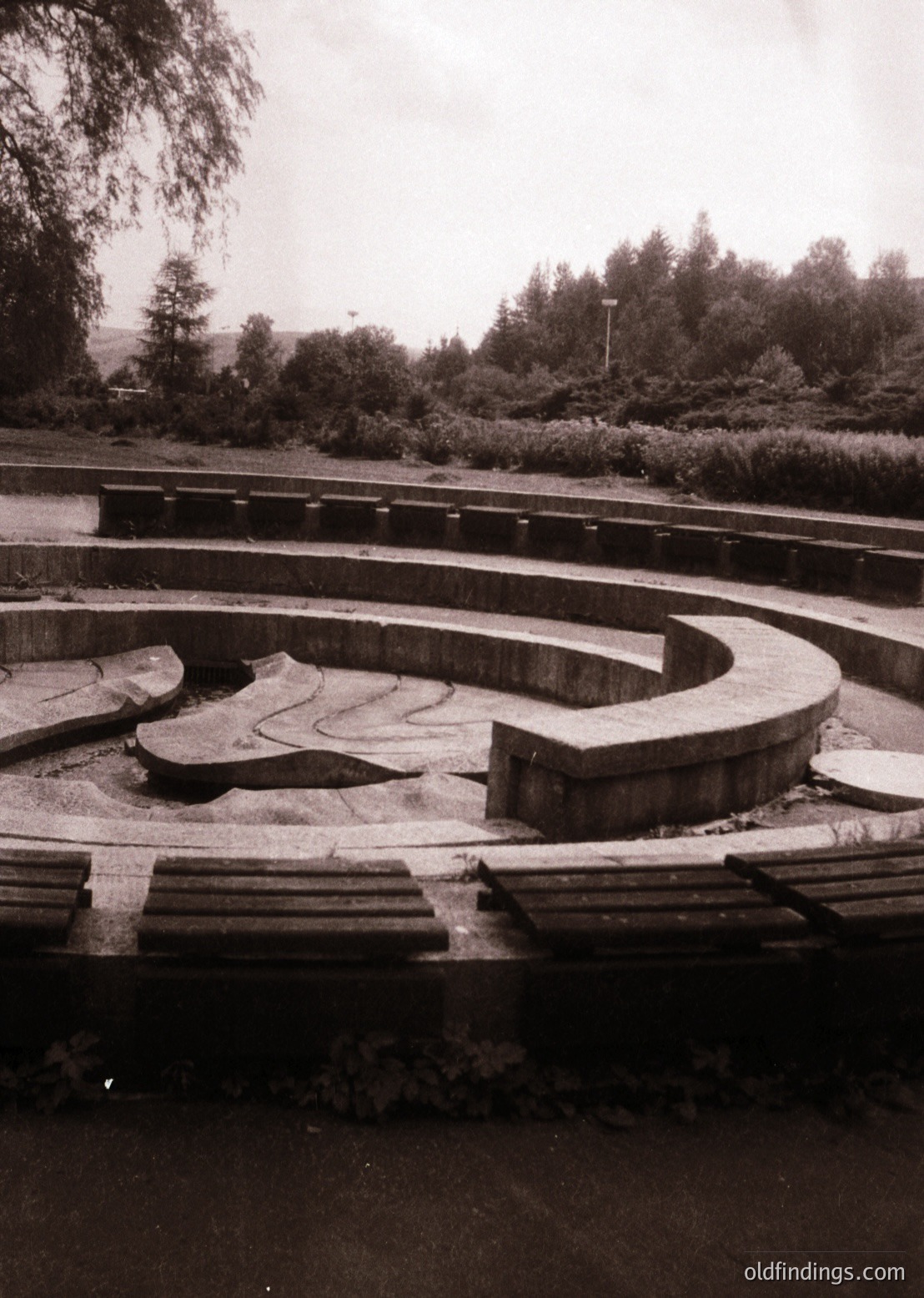 Concrete maze pathway in a landscaped park, featuring concentric loops with worn edges and overgrown vegetation. Mid-20th century urban design, likely for recreational or therapeutic use.