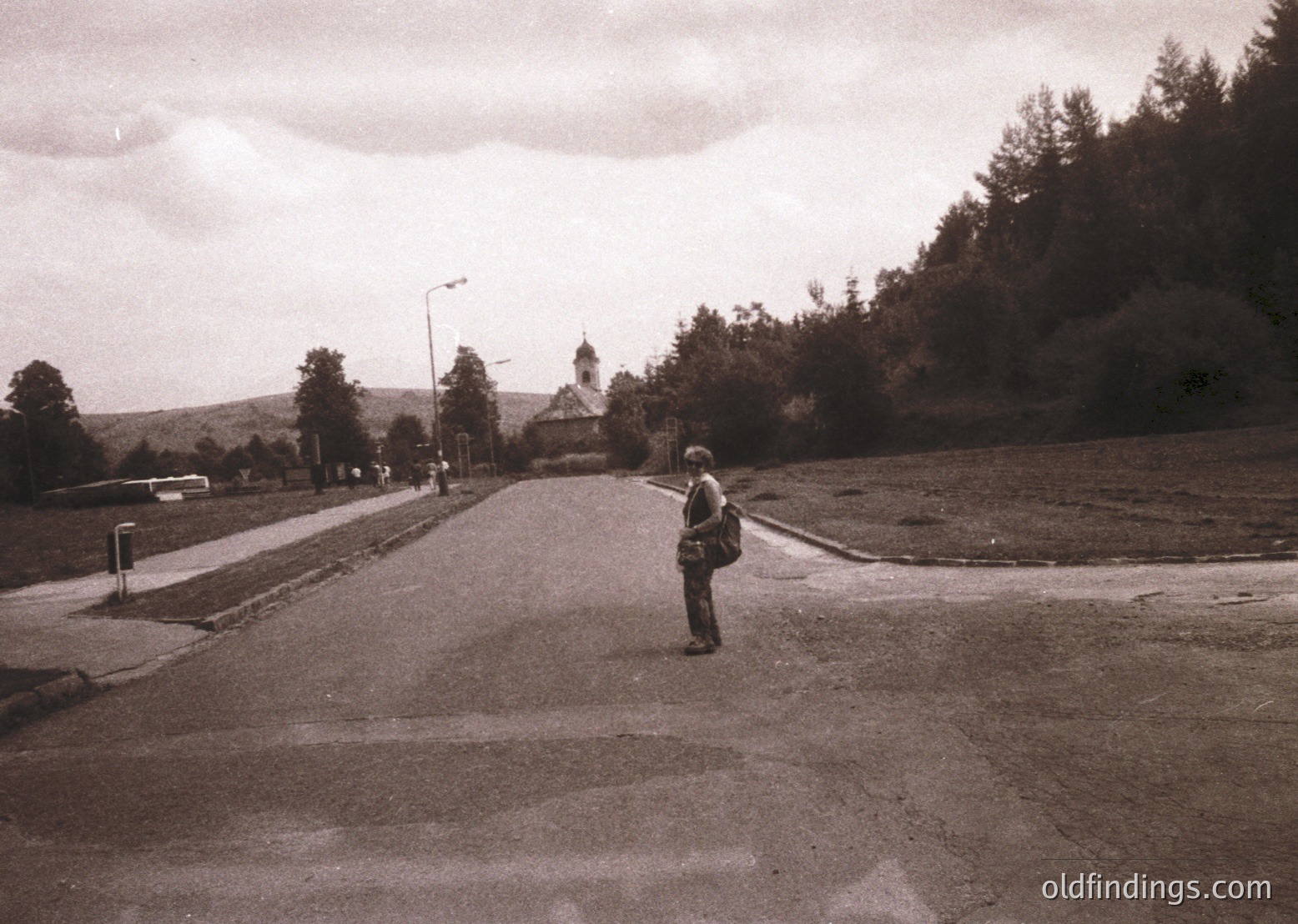 Black-and-white street scene featuring a lone pedestrian in a wide-brimmed hat and dark coat, walking toward a distant church with a bell tower. Surrounding greenery and sparse traffic suggest a quiet, possibly rural or suburban setting. Mid-20th century (1950s–1960s) architecture and clothing hint at post-war European design.