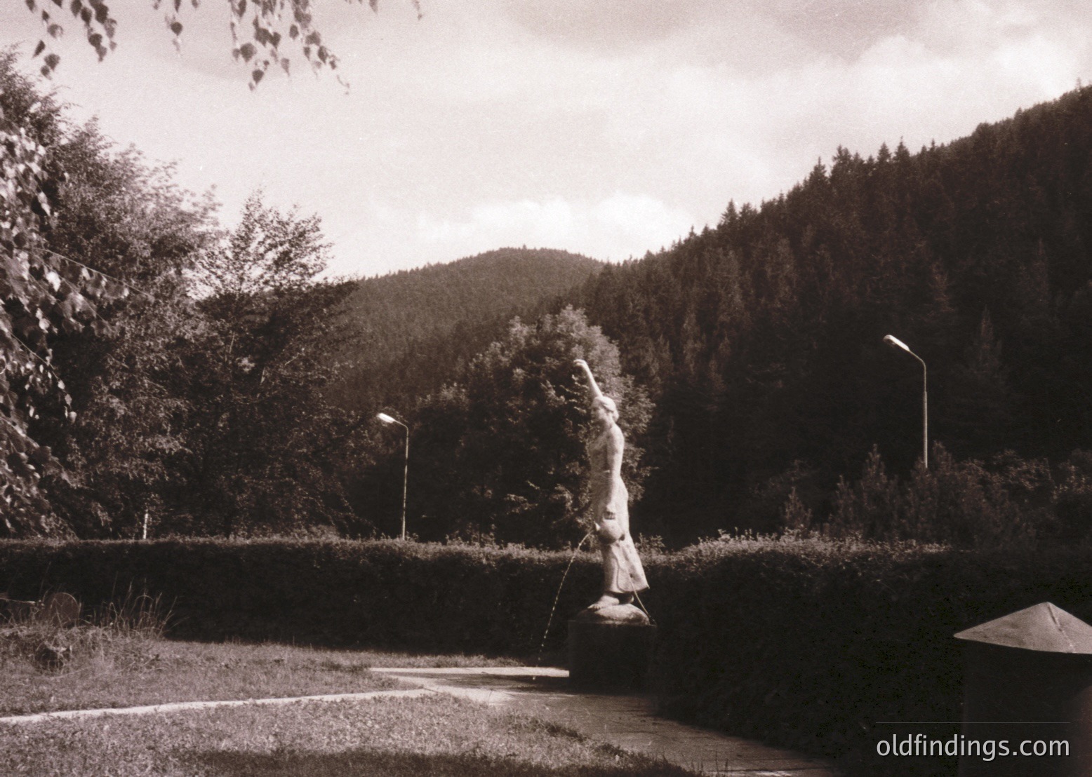 Black-and-white street scene featuring a lone figure casting a fishing line into a shallow riverbed, framed by dense foliage and a forested hillside. Concrete curb and roadside lamp post suggest urban or suburban setting. Likely 1960s–1980s vintage.