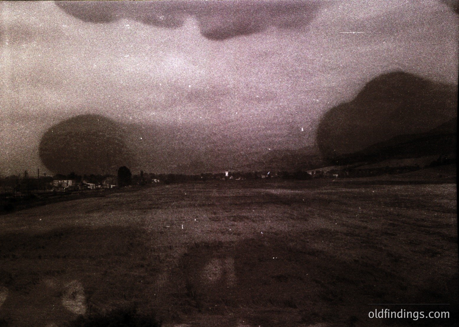 Vintage sepia-toned aerial view of a rural landscape with dense cloud cover. Low-lying fields and scattered buildings suggest agricultural activity. Likely 20th-century farming region, possibly Eastern Europe. Ideal for historical research or nostalgic design references.