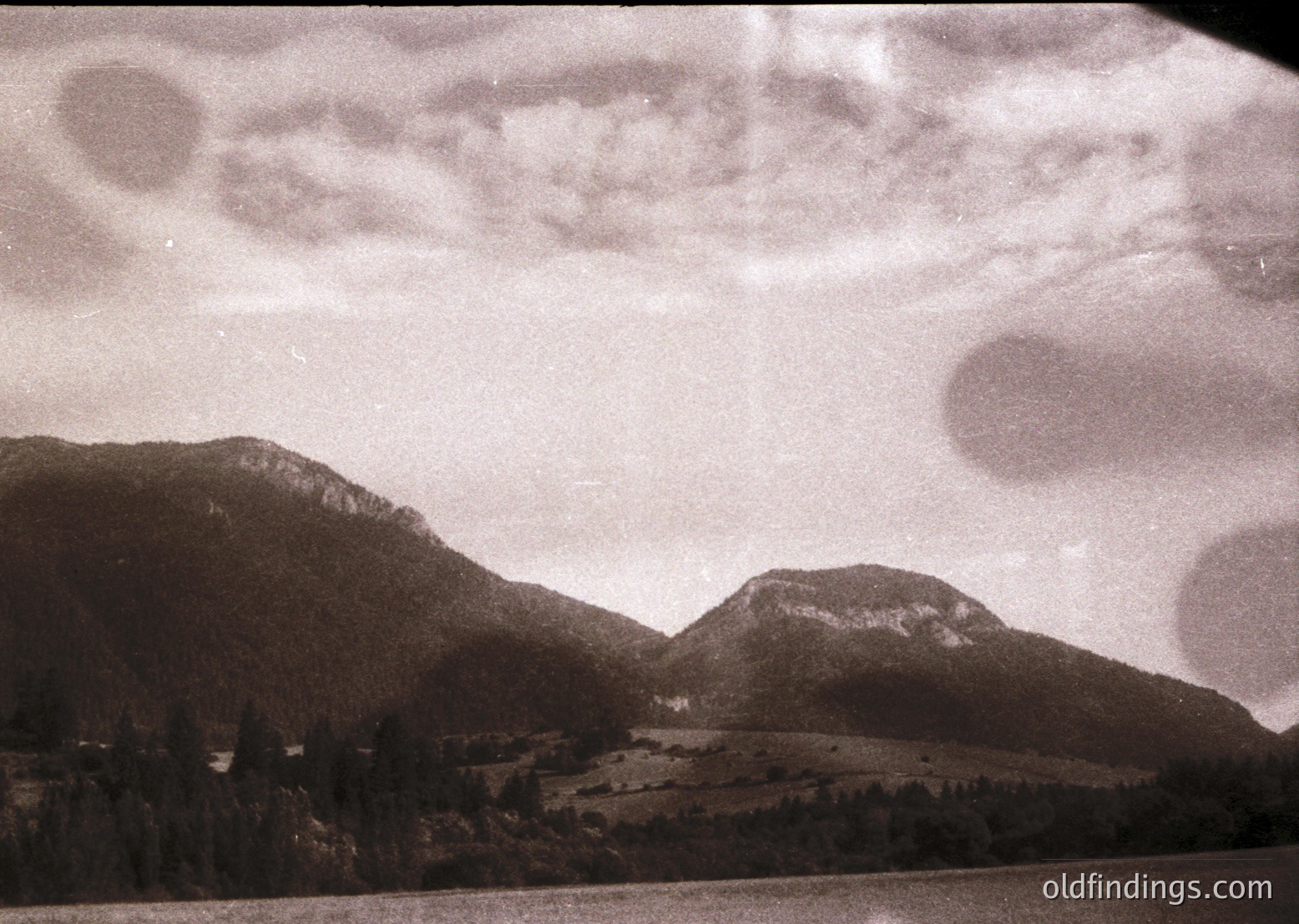 Vintage black-and-white alpine landscape with dramatic cloud formations over jagged peaks. Rolling meadows and sparse forest below. Likely 1950s–1970s European mountain region.
