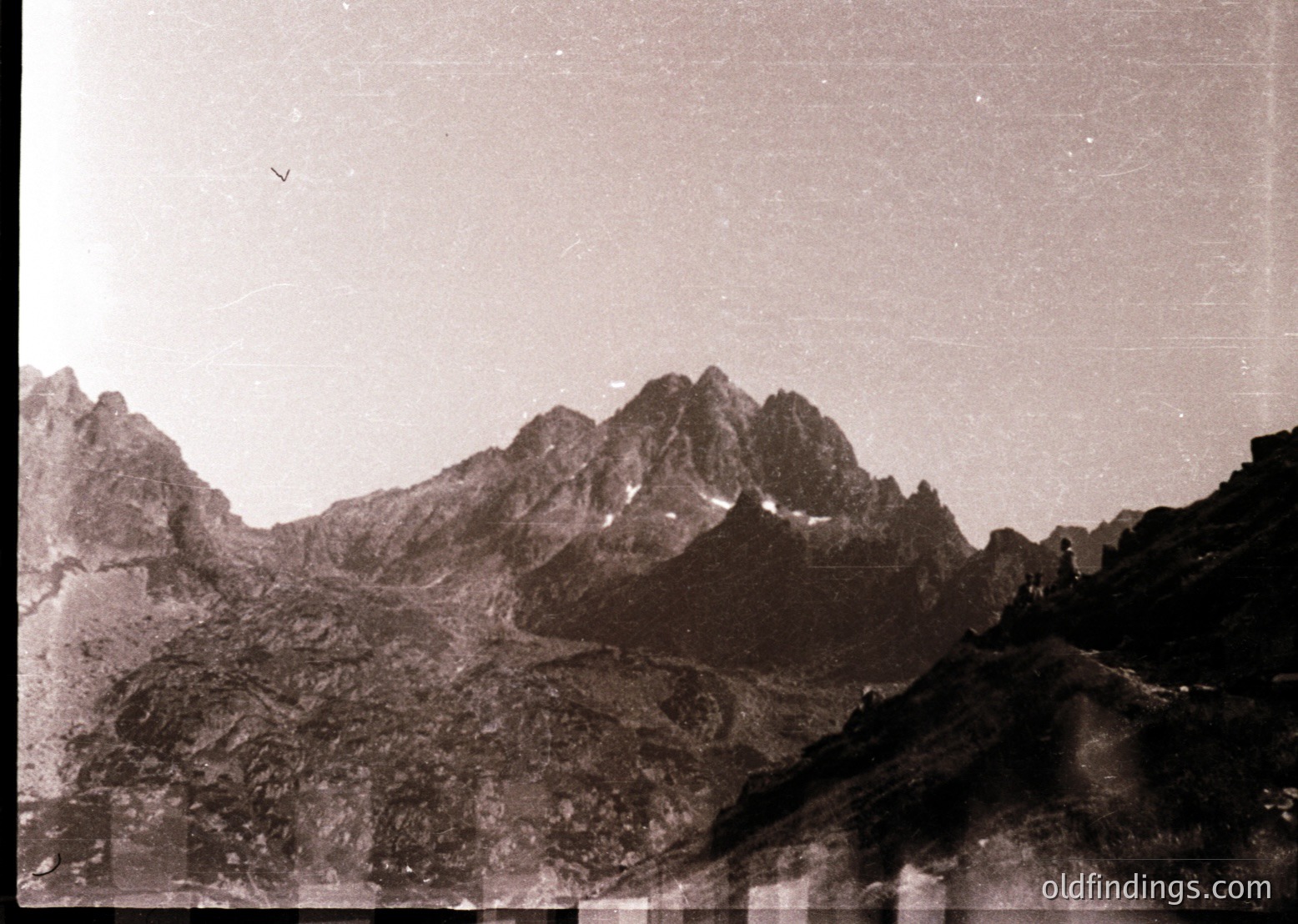 Vintage sepia-toned alpine landscape with jagged peaks and rocky terrain, likely from mid-20th century. Single solitary figure near summit, emphasizing scale.