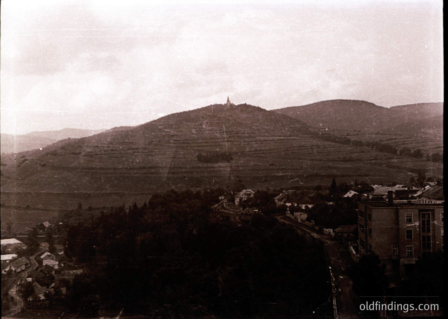 Vintage sepia-toned aerial view of a small European town nestled between terraced hills and dense forest. Prominent church spire atop a hill dominates skyline; early 20th-century architecture visible below.
