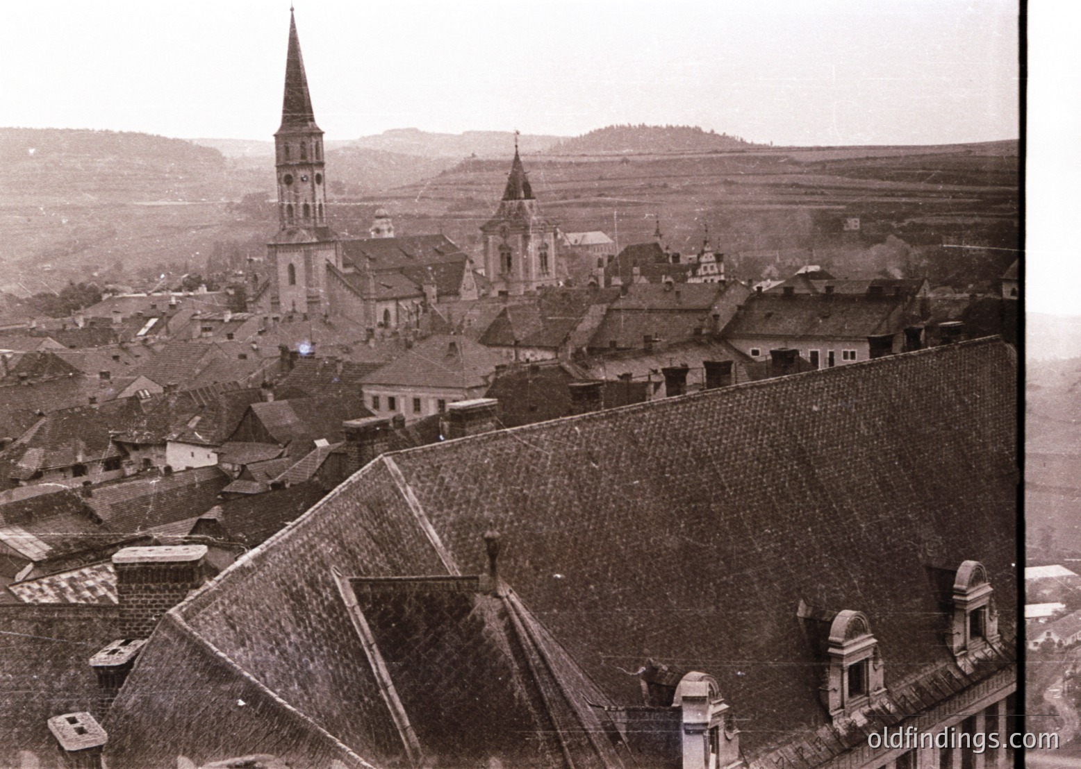 Vintage sepia-toned aerial view of a European village with distinct church spires and tiled rooftops, likely early 20th century. Prominent brick church with a tall, pointed steeple dominates the skyline. Rural landscape with rolling hills and agricultural fields in background.
