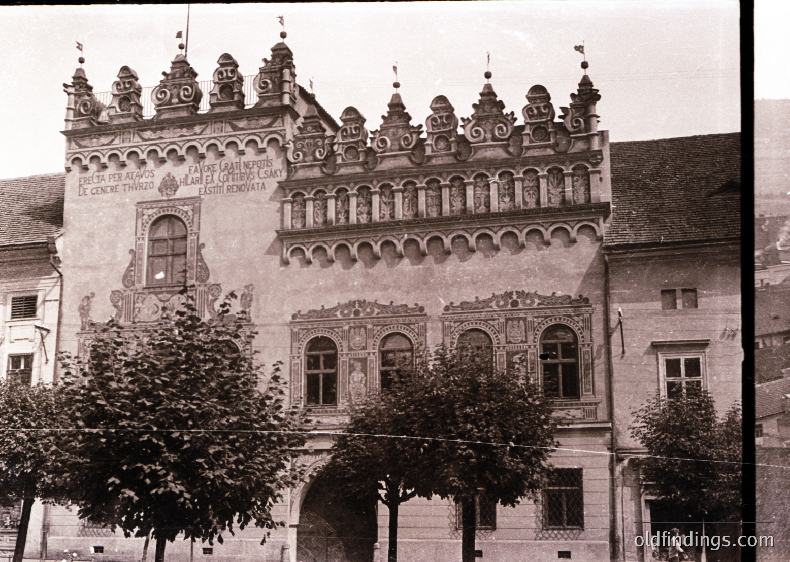 Historic Baroque-style building with ornate stonework, featuring intricate carvings, arched windows, and decorative spires. Latin inscriptions suggest cultural/historical significance, likely European. Early 20th-century sepia-toned photograph.