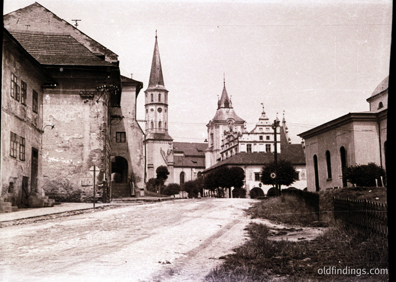 Historic European village street with a mix of Gothic and Baroque architecture. Prominent church tower with a spire and adjacent castle-like building with stepped gables. Cobblestone road and aged stone buildings suggest early-to-mid 20th century.