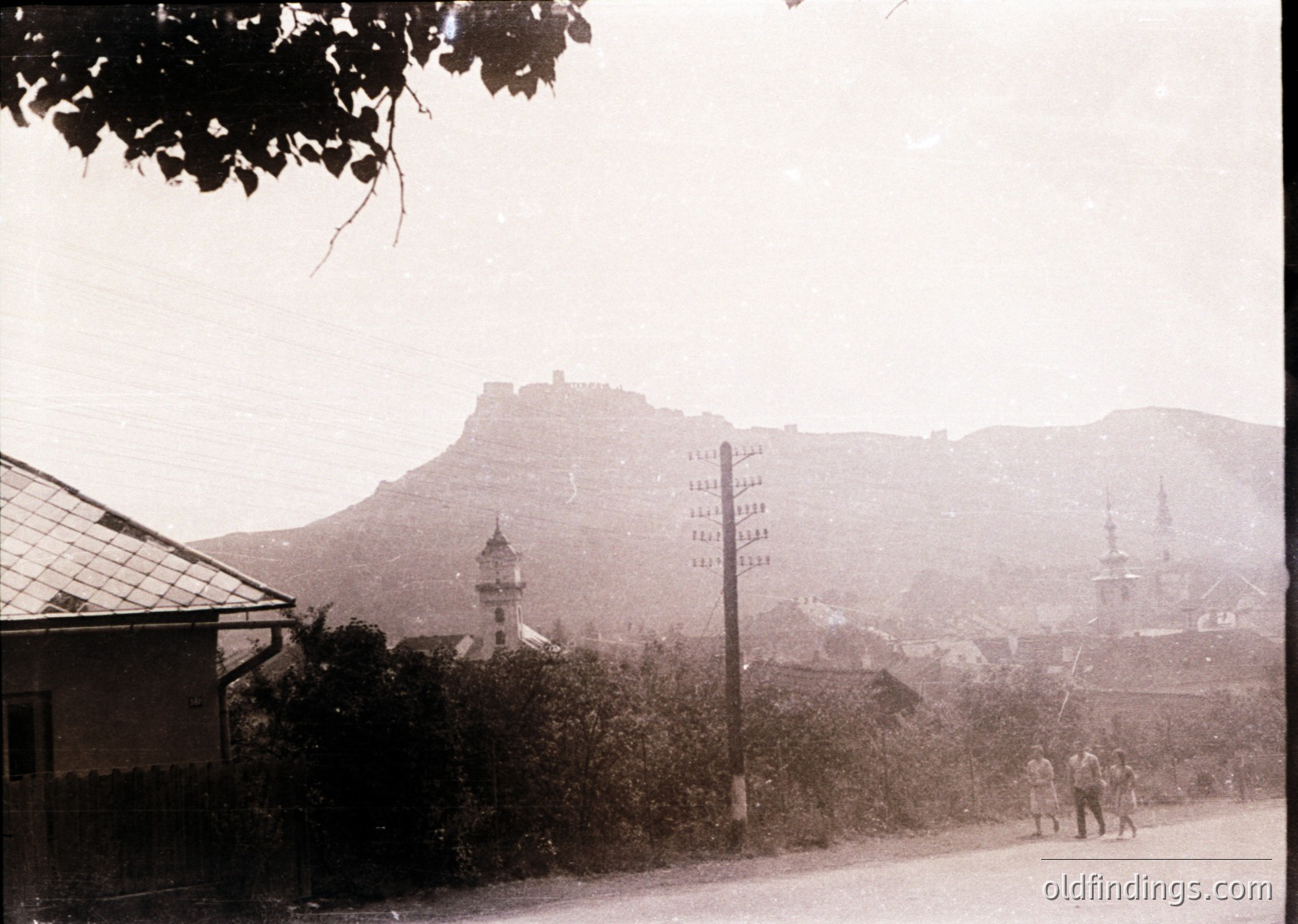 Black-and-white street scene featuring a hilltop fortress and Ottoman-era minarets in the background. Two pedestrians walk along a paved road lined with vintage street signs. Residential buildings with tiled roofs flank the left. Foggy atmosphere suggests early 20th-century Balkans, likely ’s . Ideal for historical research and vintage design references.