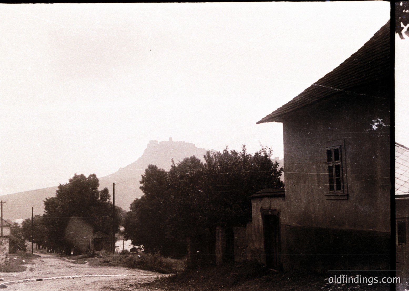 Black-and-white rural street scene with a stone house featuring wooden-framed windows and a sloped roof. A rocky hilltop fortress looms in the background, surrounded by dense trees and sparse buildings. Likely early-to-mid 20th century, Eastern European setting.