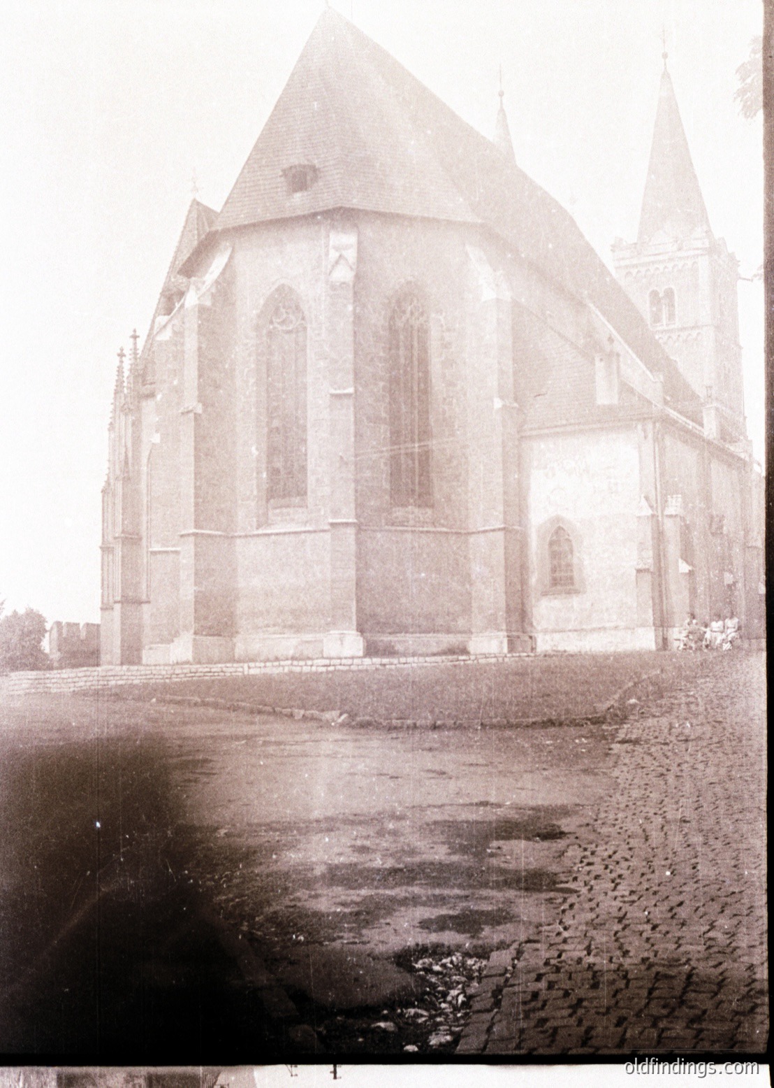 Gothic-style church with pointed arches, buttresses, and a steeply pitched roof, likely from the 12th–14th century. Wet cobblestone courtyard in foreground suggests rain or seasonal dampness. Architectural details include lancet windows and a central tower.