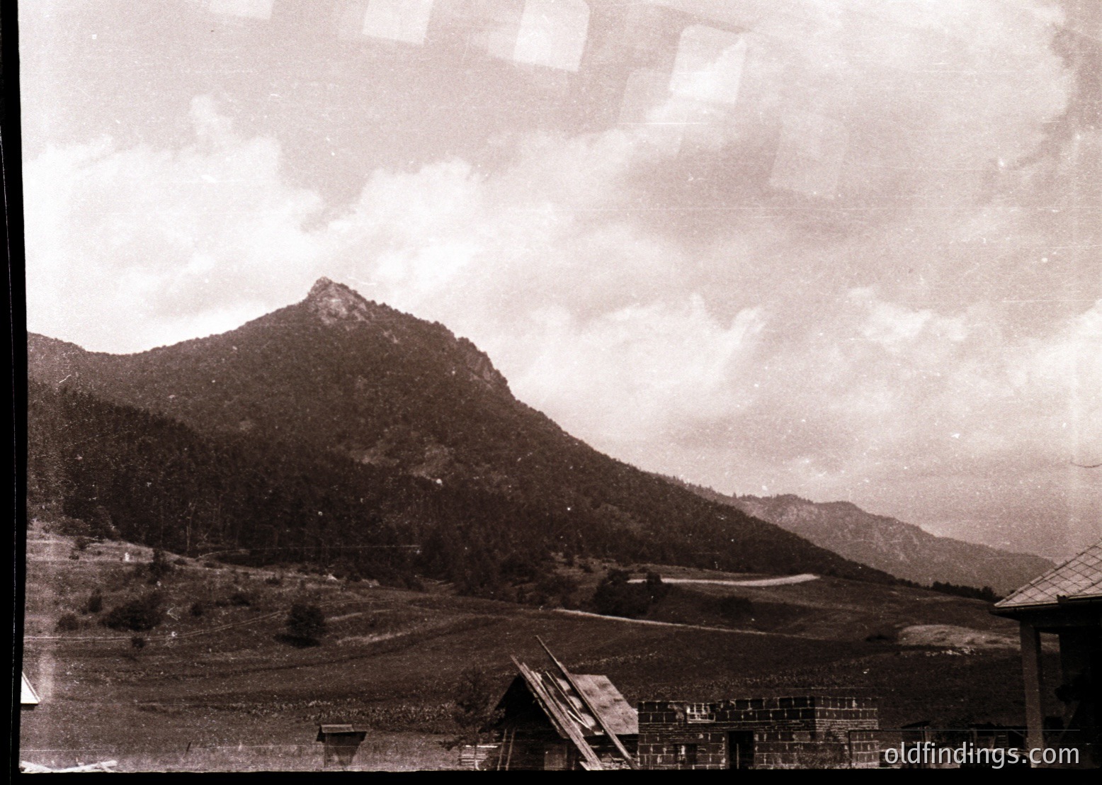 Vintage sepia-toned landscape featuring rugged mountain terrain with sparse vegetation. Foreground includes a small wooden structure and agricultural plots, likely for farming. Overcast sky enhances dramatic contrast. Suggests rural, possibly alpine setting from mid-20th century.