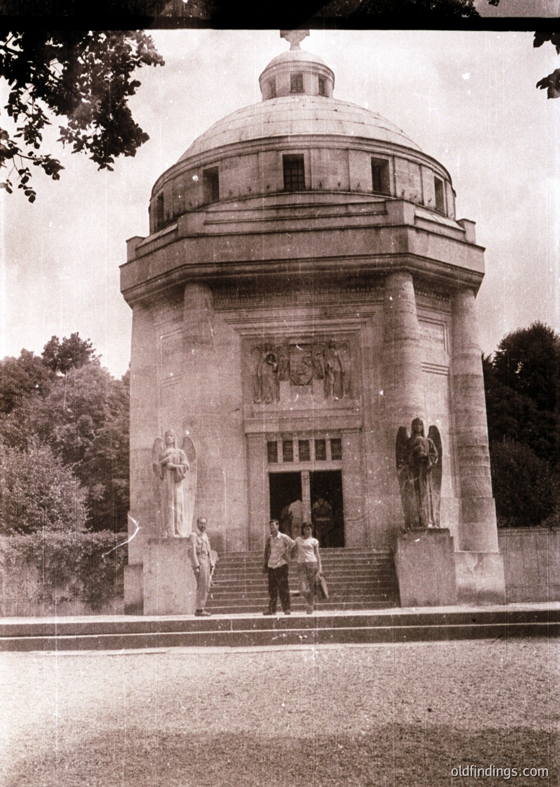 Neoclassical mausoleum with domed roof, flanked by sculpted figures and reliefs depicting horses. Three men in 1930s-era suits pose on stone steps. Lush greenery and mature trees frame the structure, suggesting a park or memorial setting.
