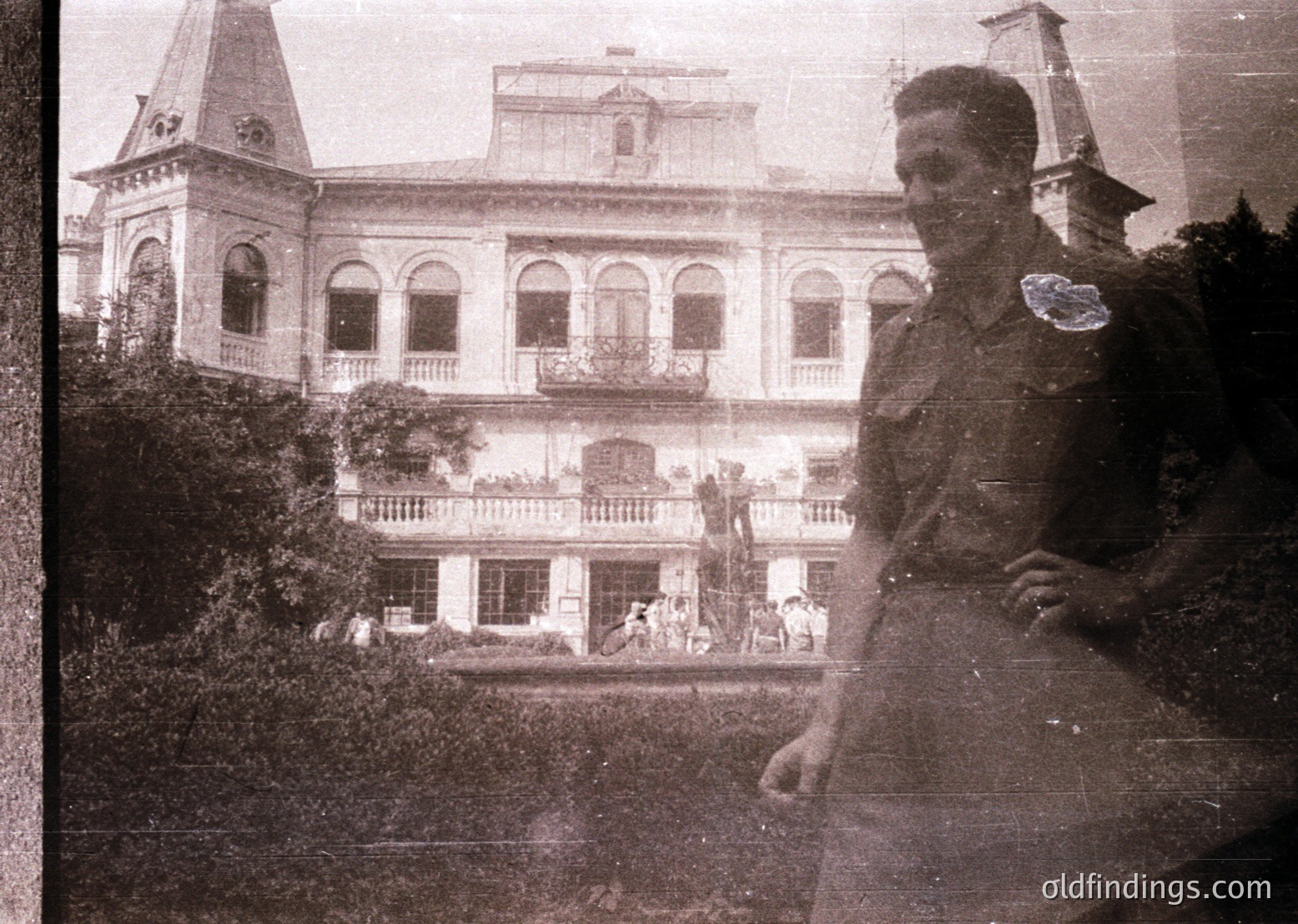 Vintage sepia-toned photo of a grand colonial-era building with arched balconies, central tower, and lush greenery. Uniformed man in foreground suggests military or official presence, likely mid-20th century. Crowd on balconies and courtyard hints at social gathering or event.