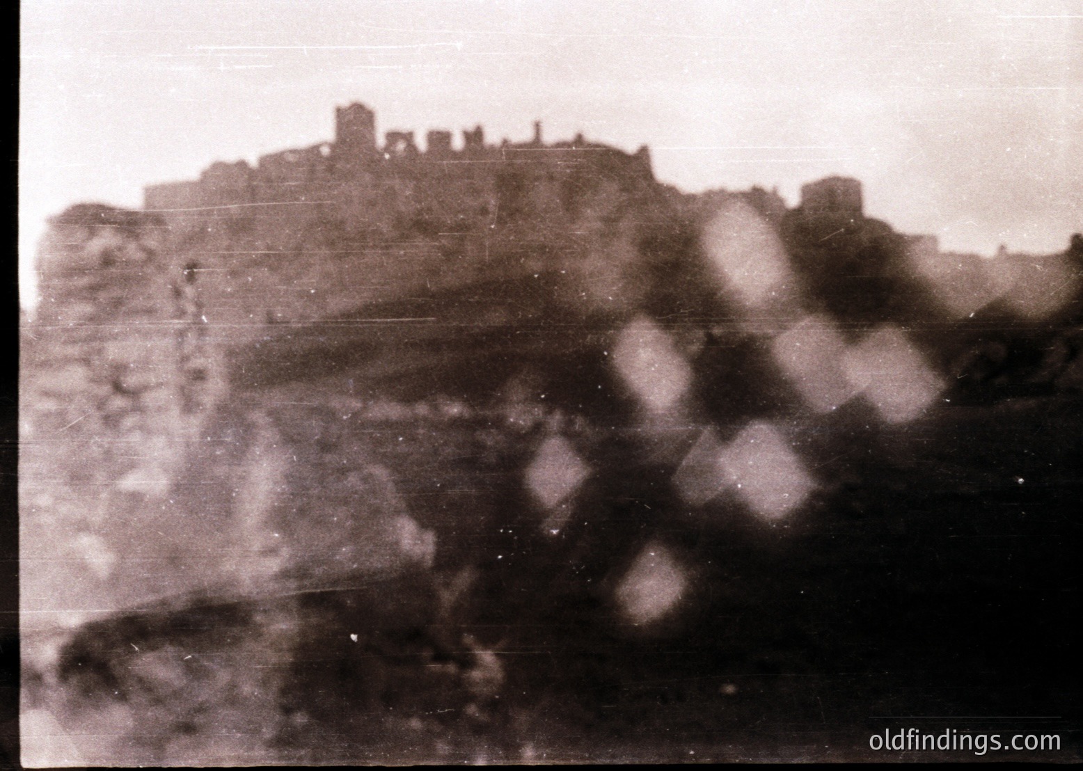 Vintage sepia-toned photo of a hilltop fortress with crumbling stone walls and towers, likely Eastern European. Fog obscures lower details, emphasizing atmospheric mystery. Mid-20th century architectural ruins suggest historical significance.
