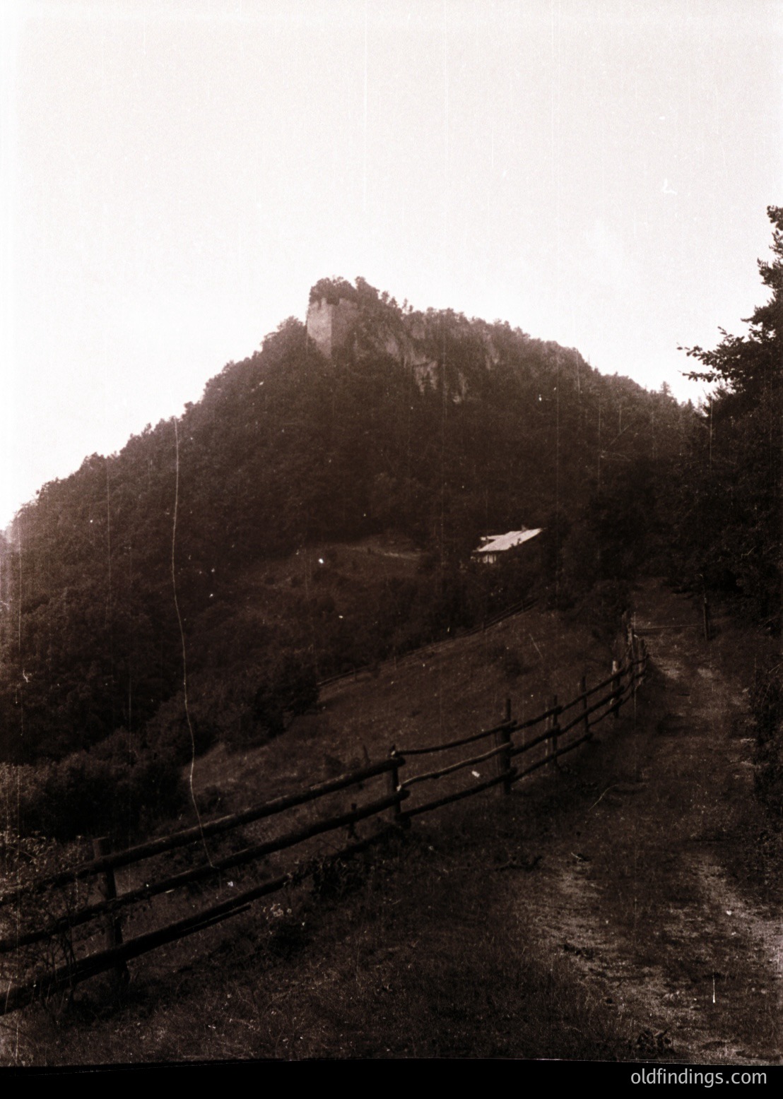 Black-and-white sepia-toned mountain landscape with a winding wooden fence leading to a rocky summit. Mist obscures the peak, creating a dramatic, atmospheric scene. Likely early-to-mid 20th century due to photographic style.
