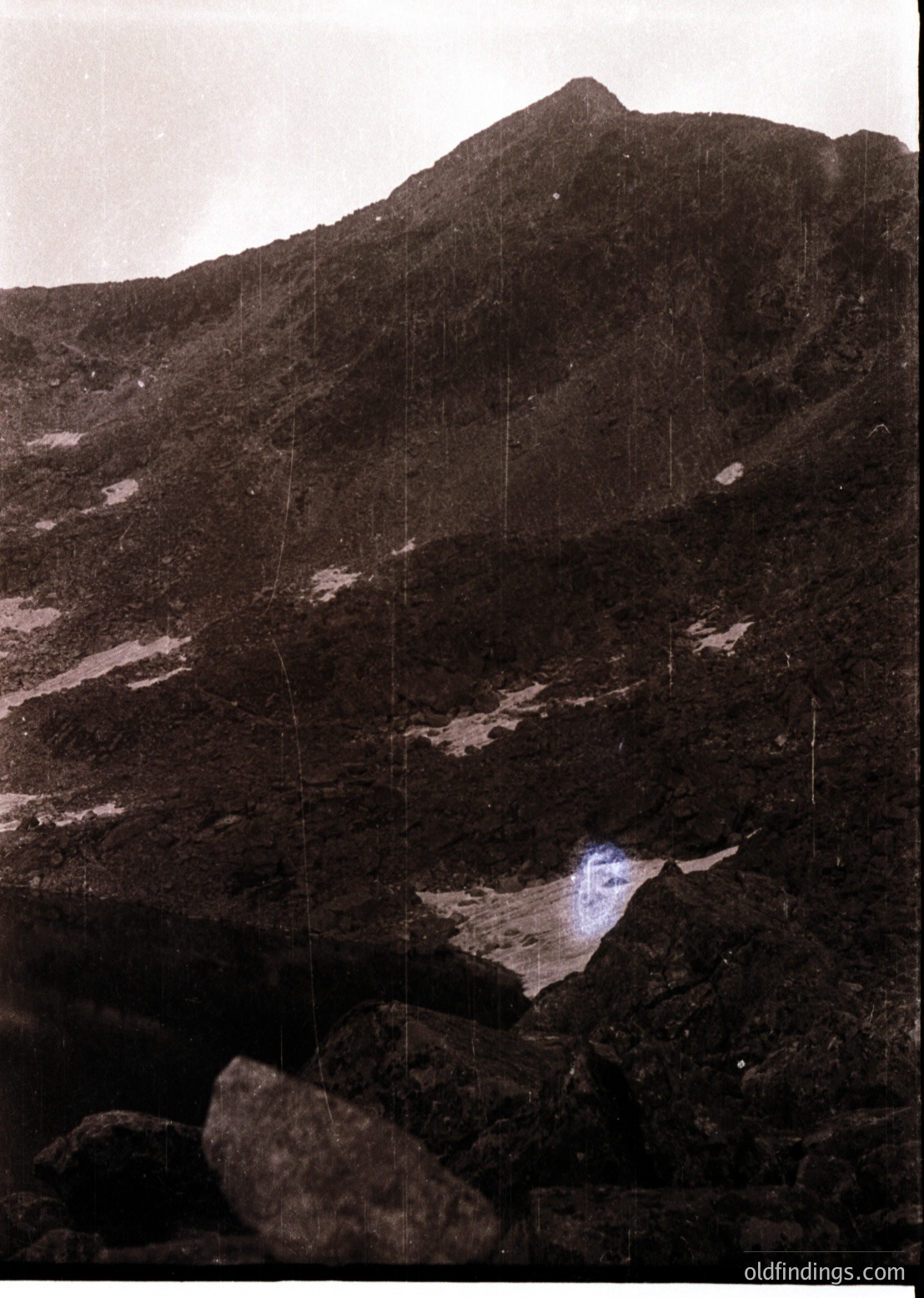 Black-and-white alpine landscape featuring jagged rock formations and a small, reflective water body in a narrow valley. Vertical light streaks suggest long-exposure photography. Likely captured in the mid-20th century.