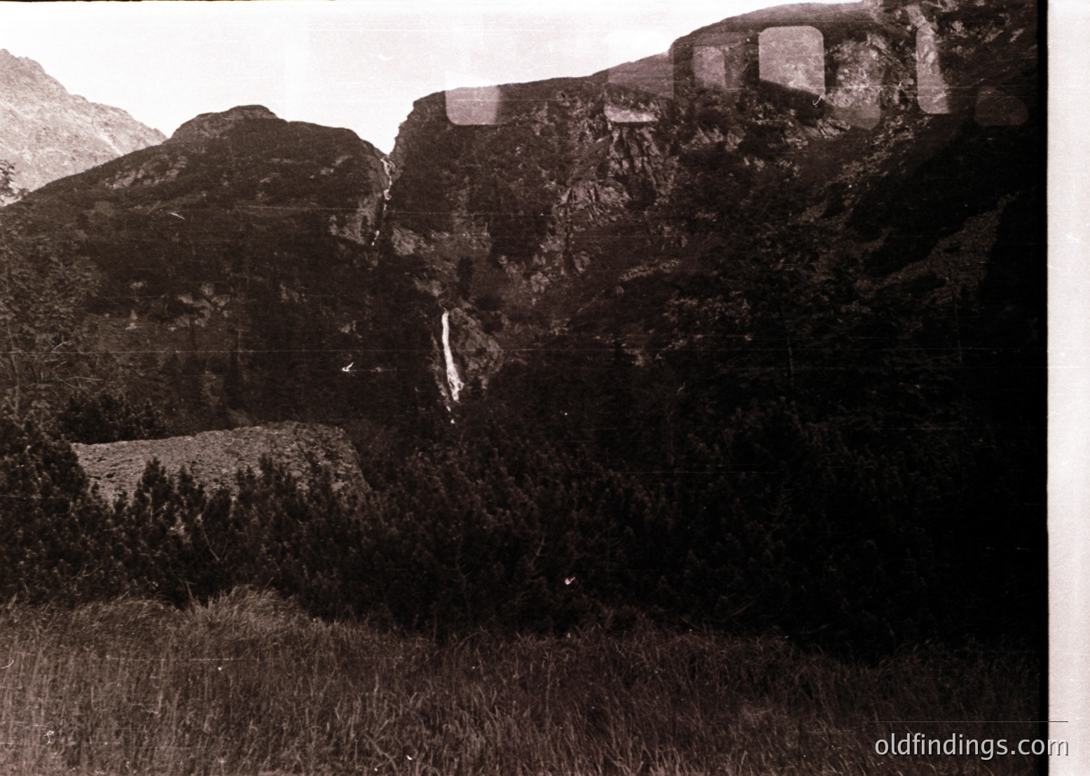 Vintage sepia-toned alpine landscape featuring jagged rock formations and a cascading waterfall. Dense forest covers lower slopes, with sparse grassy meadows in the foreground. Likely early 20th-century European mountain region.