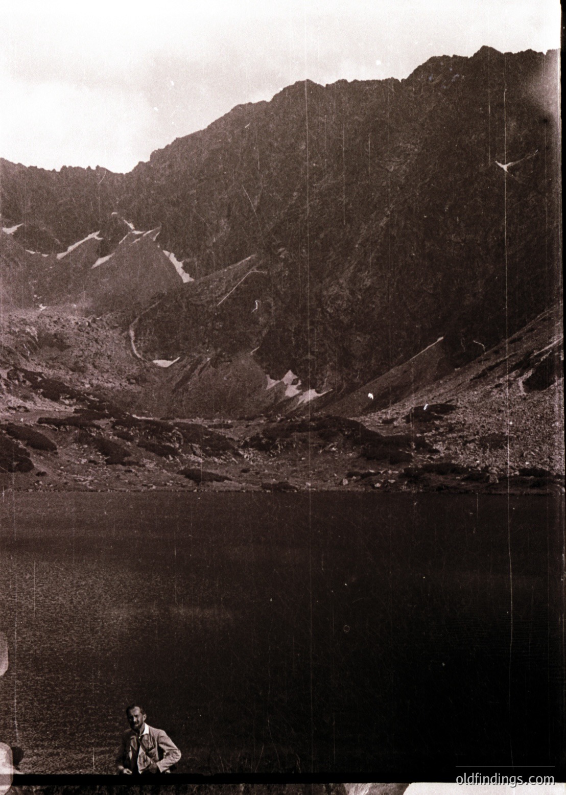 Mid-20th century black-and-white alpine lake scene with rugged peaks. A lone figure in outdoor gear sits by the water’s edge, framed by steep, snow-dusted slopes. Classic vintage photography style suggests or European Alps.