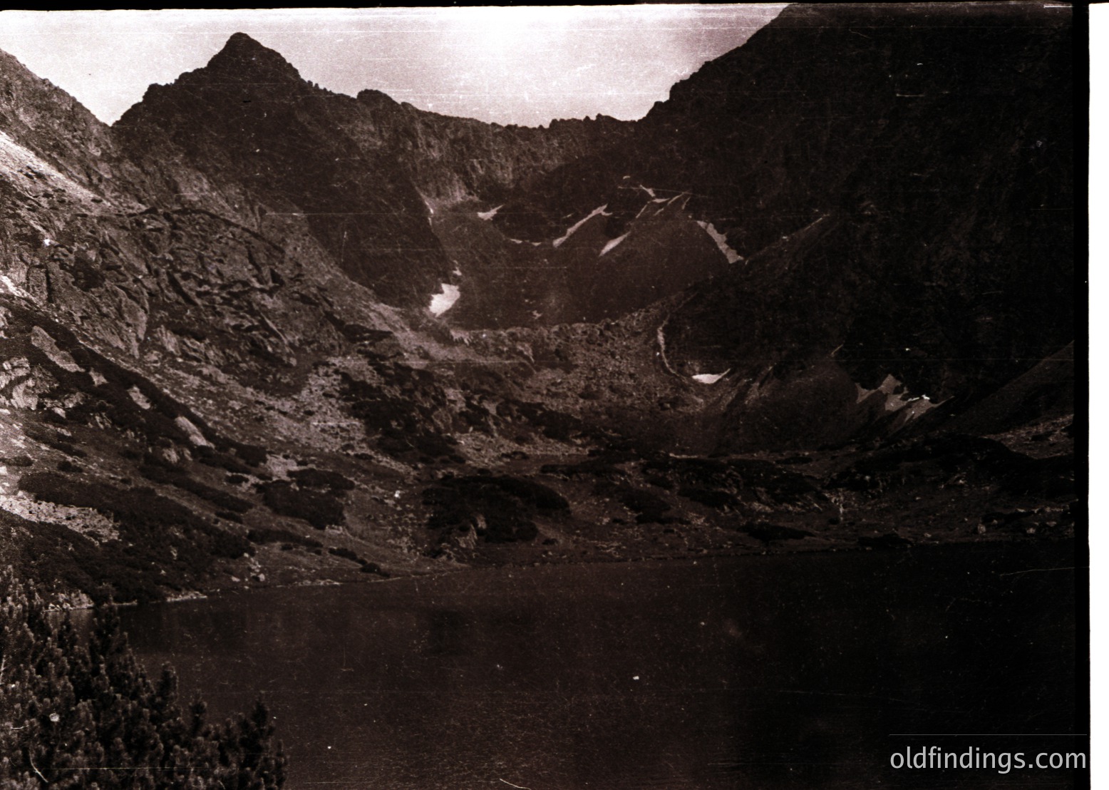 Black-and-white aerial view of a rugged alpine valley with steep, forested slopes and a winding river cutting through rocky terrain. Snow patches highlight elevation changes. Likely mid-20th century due to monochrome and composition style.