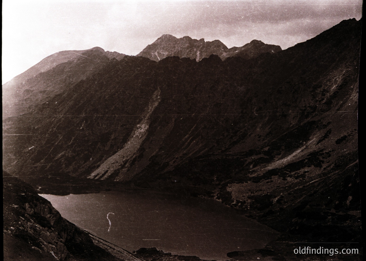 Black-and-white alpine lake nestled between jagged, forested ridges under overcast skies. Dramatic rock formations frame serene water, suggesting remote high-altitude terrain. Likely early-to-mid 20th century due to sepia tone and composition style.