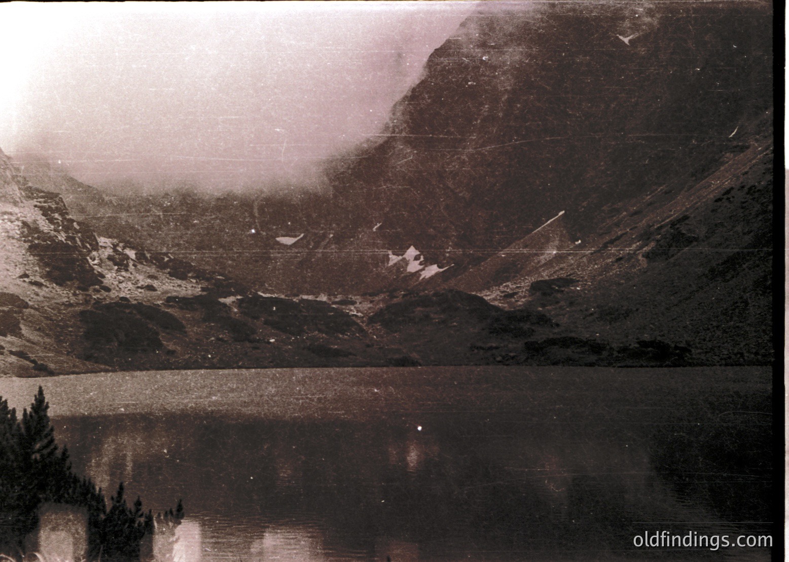 Vintage sepia-toned alpine lake framed by rugged, forested slopes and misty peaks. Reflections mirror the misty terrain in calm waters. Likely captured in the mid-20th century, possibly for travel or nature documentation.
