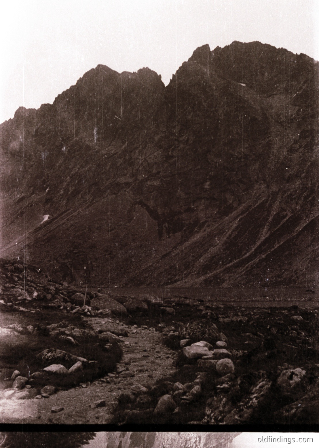 Black-and-white alpine landscape featuring jagged, snow-capped rock formations and a rocky riverbed. Vertical poles suggest early mountaineering or surveying activity. Likely late 19th to early 20th century.