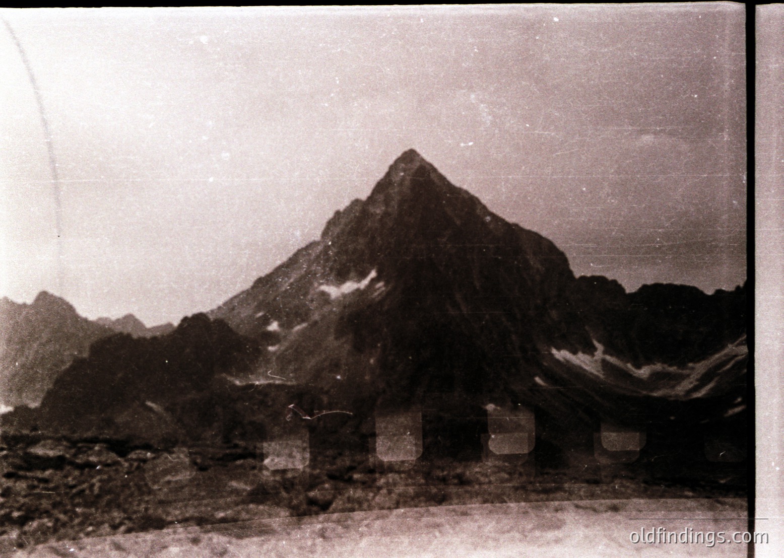 Vintage black-and-white alpine landscape featuring jagged peaks and a rocky foreground. Small stone markers or cairns align near the base, suggesting a hiking trail or summit path. Overcast skies enhance the dramatic, rugged terrain. Likely early-to-mid 20th century due to photographic style.